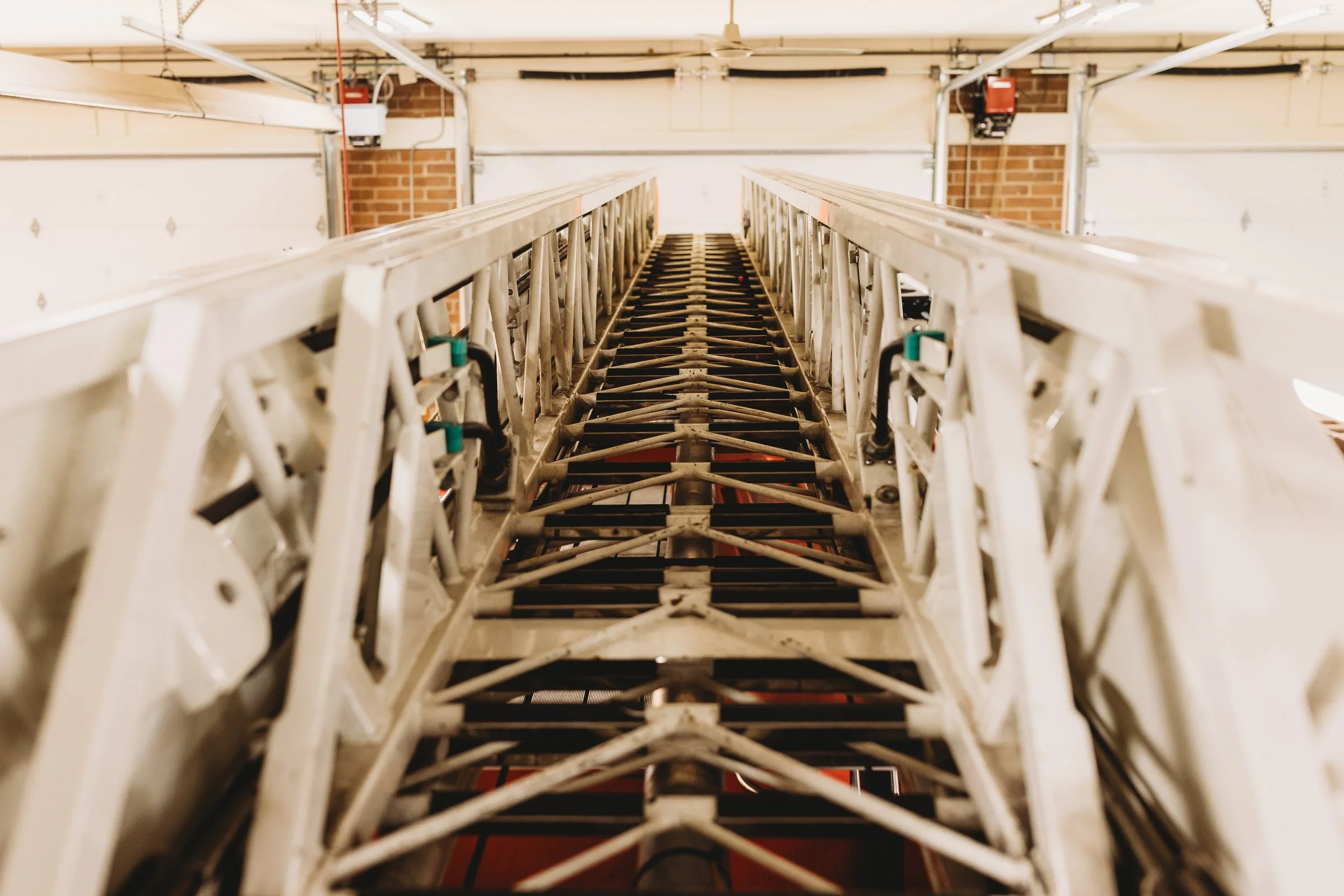 Indoor ladder with metal rungs and side rails, extending upward toward the ceiling in a garage with brick and white walls.