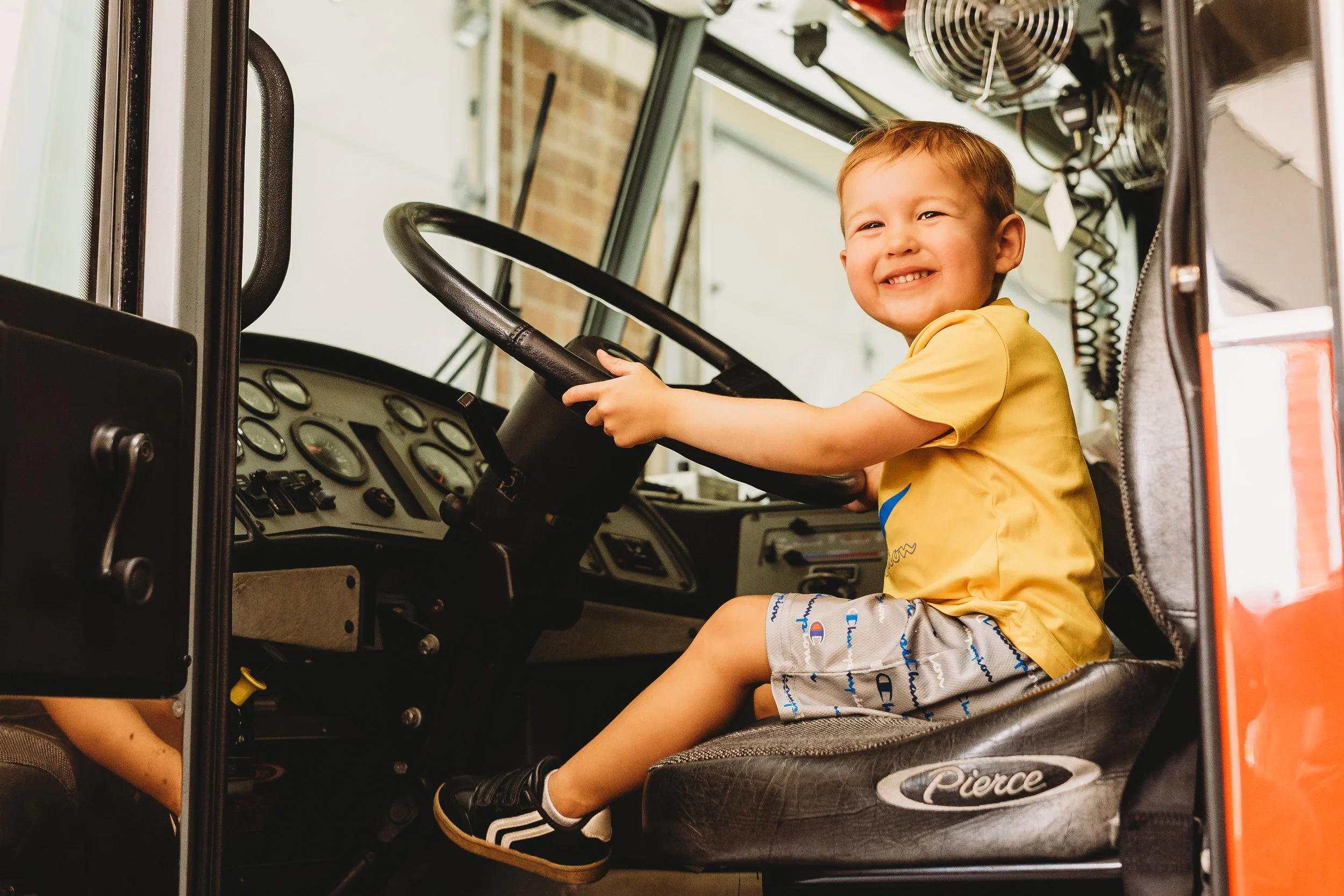 A young boy sitting in a school bus driver's seat, smiling and holding the steering wheel.