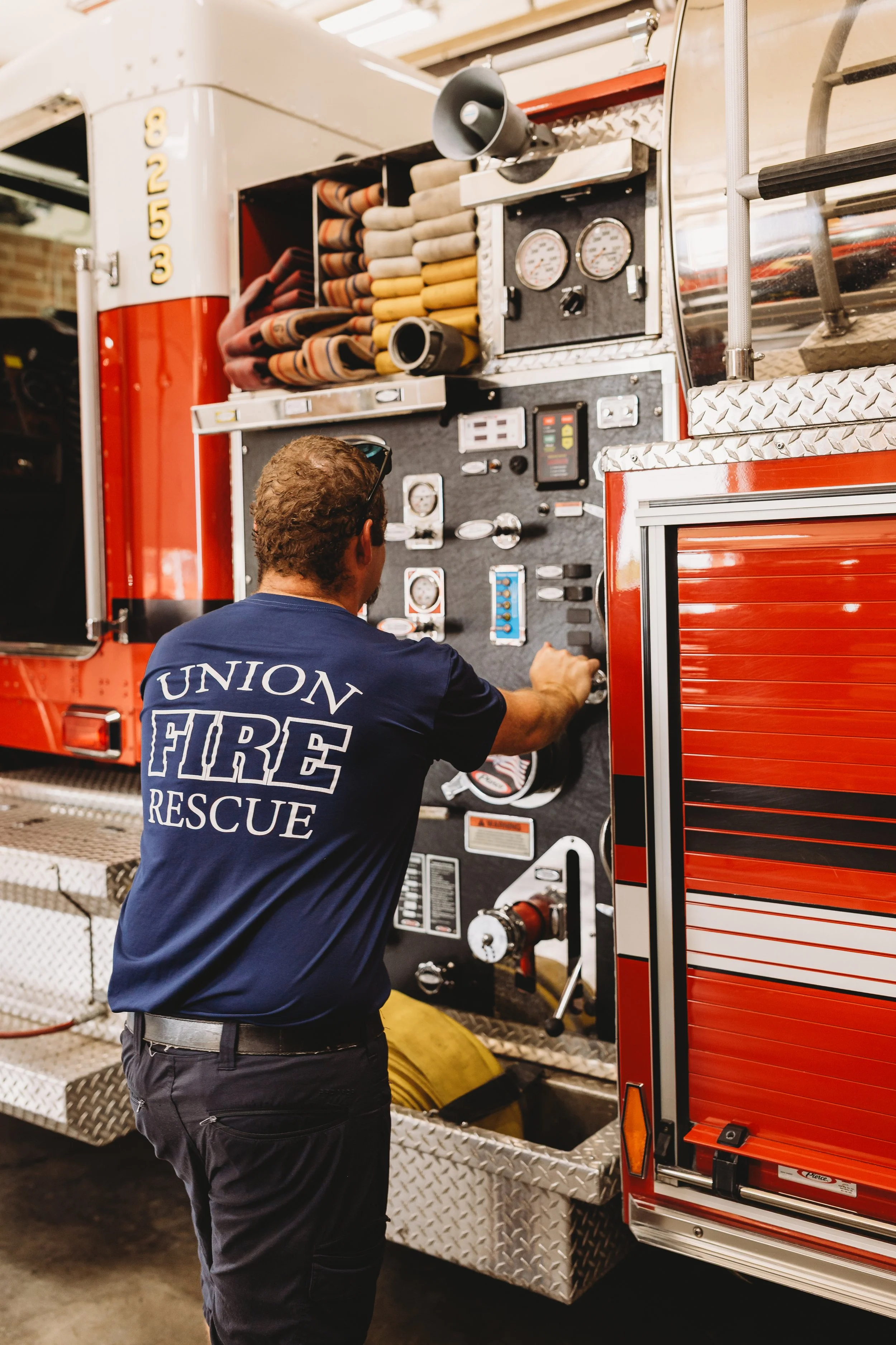 A firefighter in a navy blue shirt with 'Union Fire Rescue' on the back adjusts controls on a red fire truck.