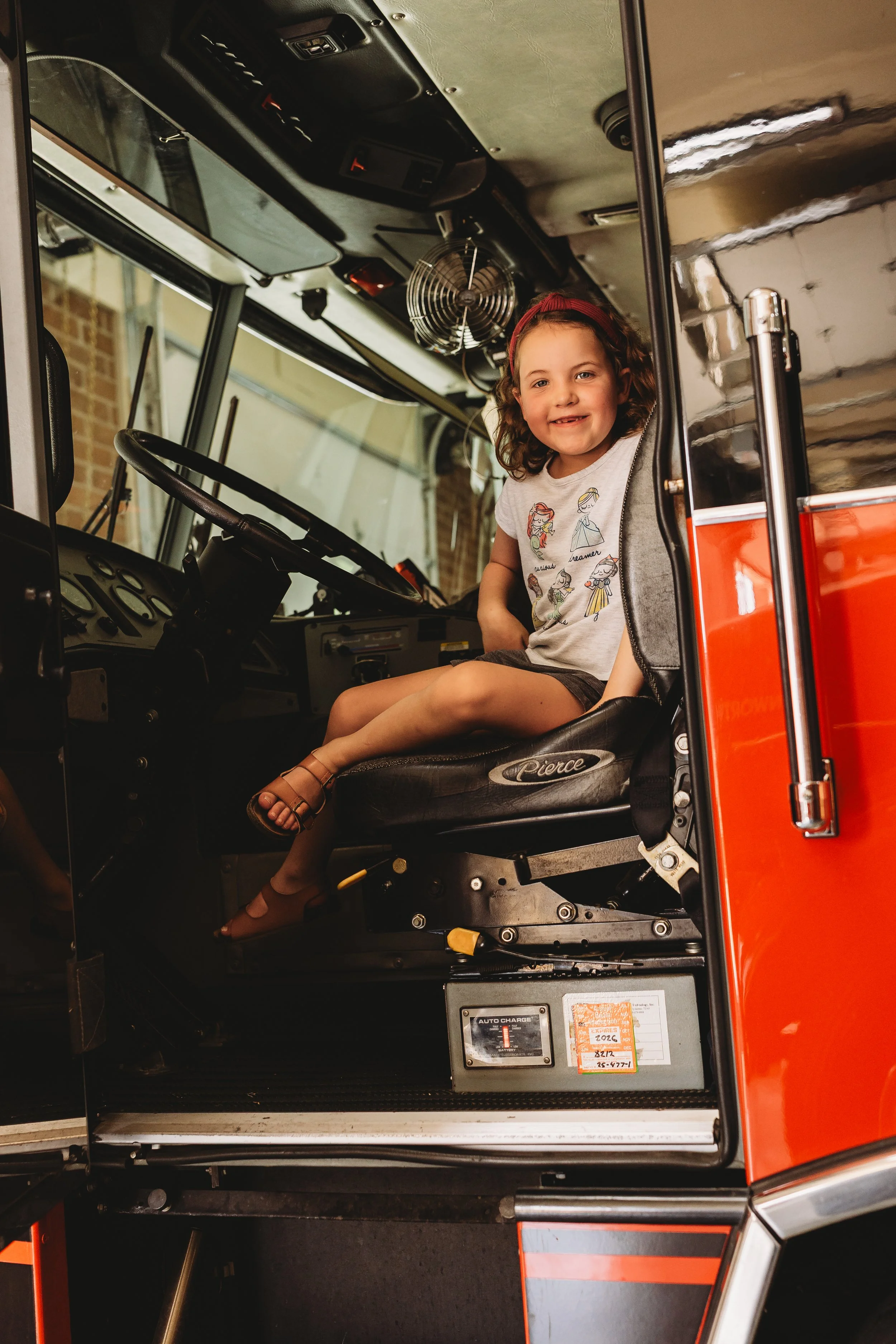 A young girl with brown curly hair, wearing a white t-shirt with colorful drawings, sitting in the driver's seat of a fire truck, smiling at the camera.
