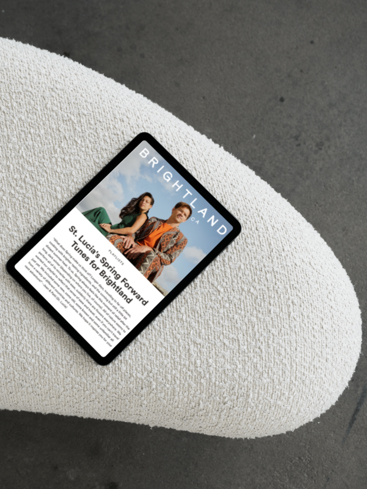 A digital tablet displaying a C__LOUISE press clip for the brand Brightland placed on a textured, off-white, curved surface. The cover features two models and an article headline about St. Lucia.