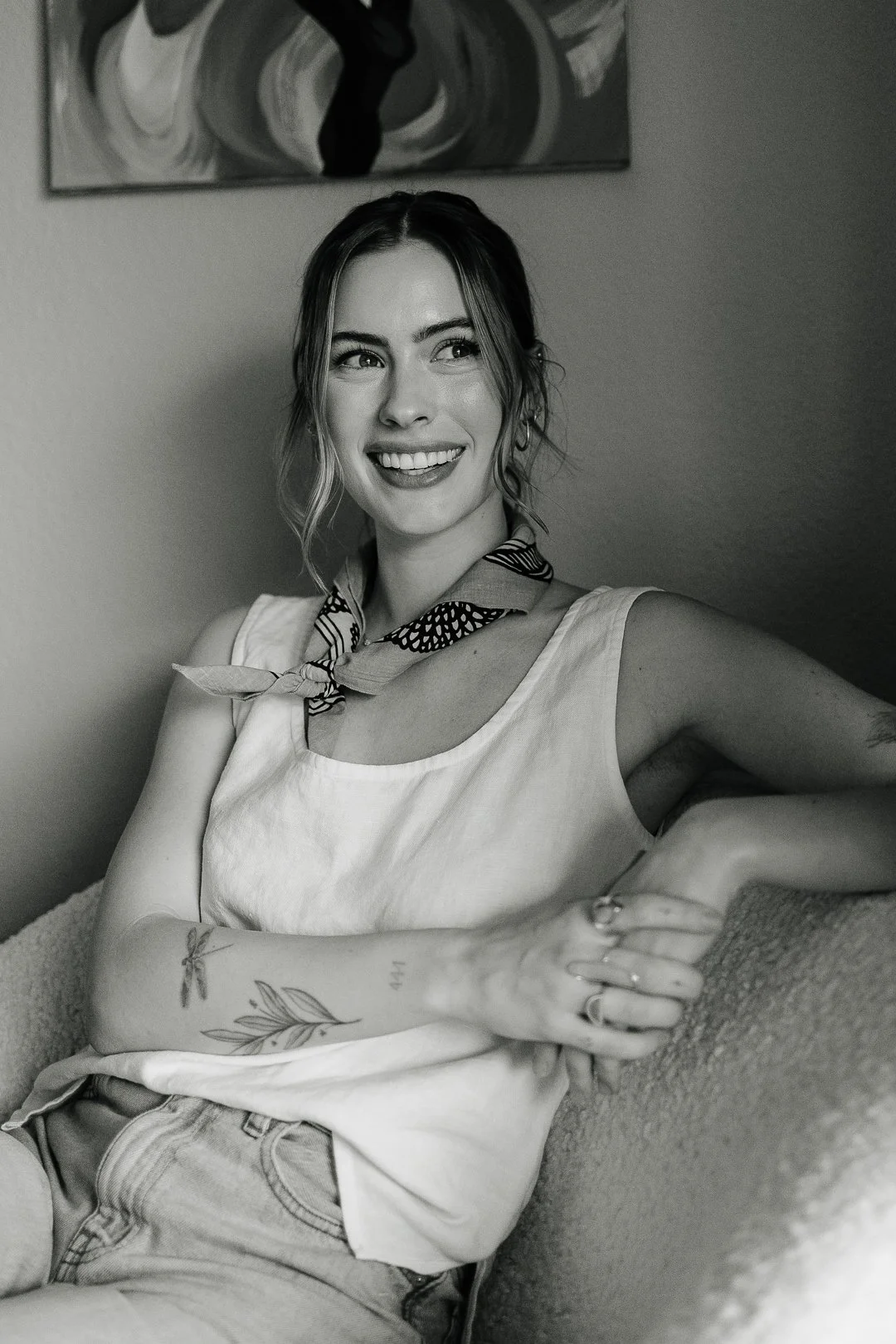 A young woman smiling and sitting on a sofa, wearing a sleeveless top and a scarf around her neck, with a tattoo on her arm and rings on her fingers.