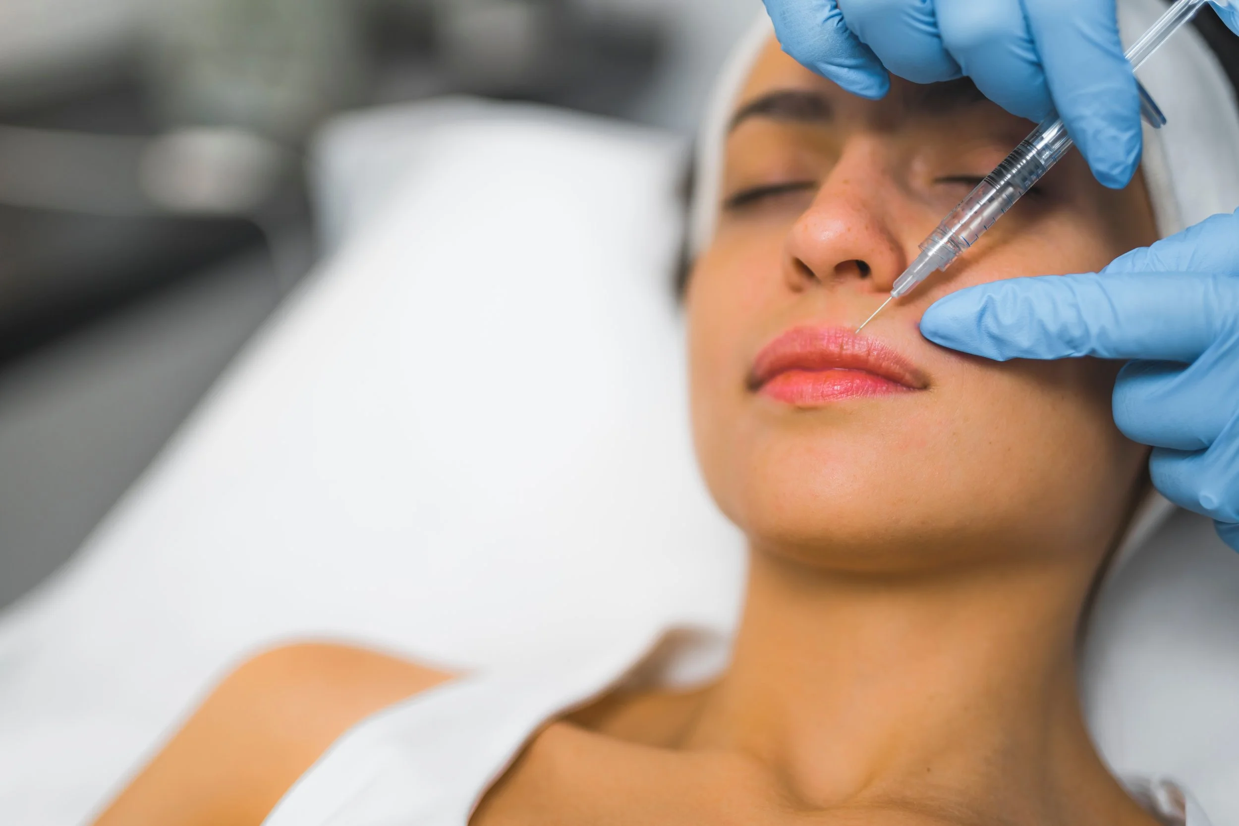 A woman receiving a cosmetic injection in her lips from a medical professional wearing blue gloves.