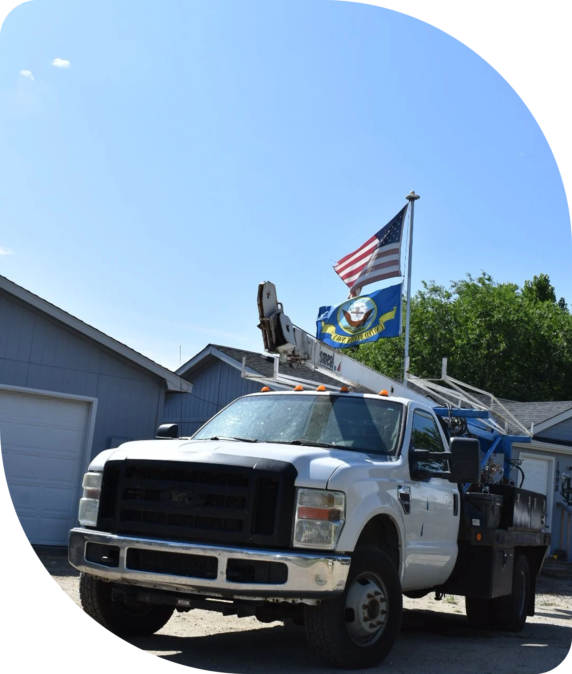 A white utility truck with a cherry picker lift, carrying two flags. The flag on the left is the United States flag, and the flag on the right appears to be a state or organizational flag with yellow, blue, and white colors. The truck is parked in front of a residential building with a garage, and there are green trees and a clear blue sky in the background.