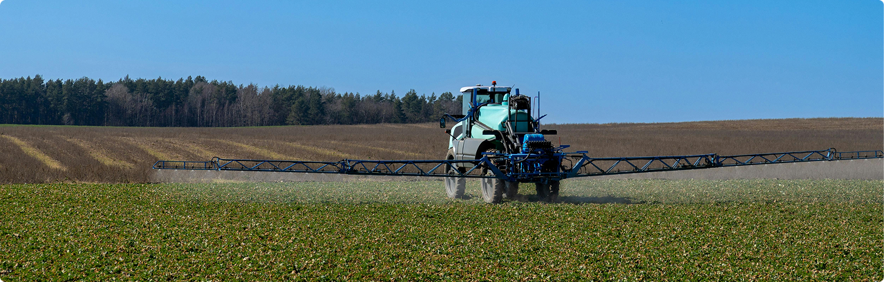 A tractor spraying crops in a field with trees in the background under a blue sky.
