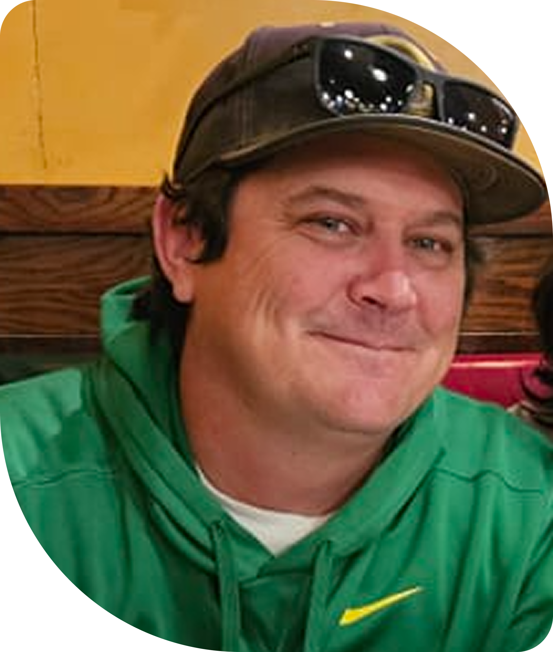 A man wearing a green hoodie and a baseball cap with sunglasses on top, sitting against a wooden background.