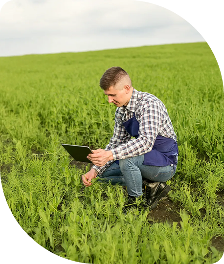 Young man crouching in a green field holding a tablet, with a cloudy sky overhead.