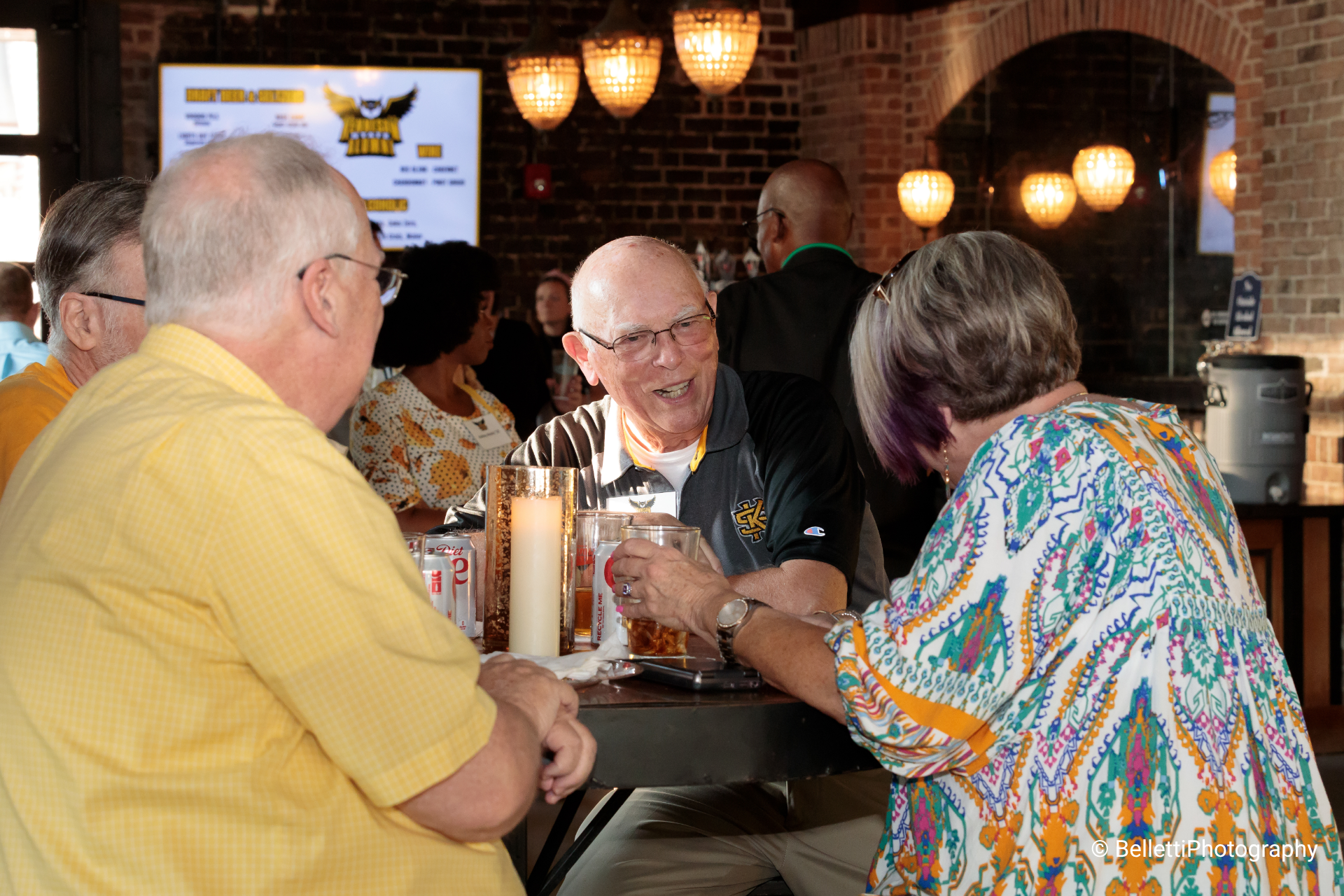 group of three elderly people seated at a high top drinking beers and chatting