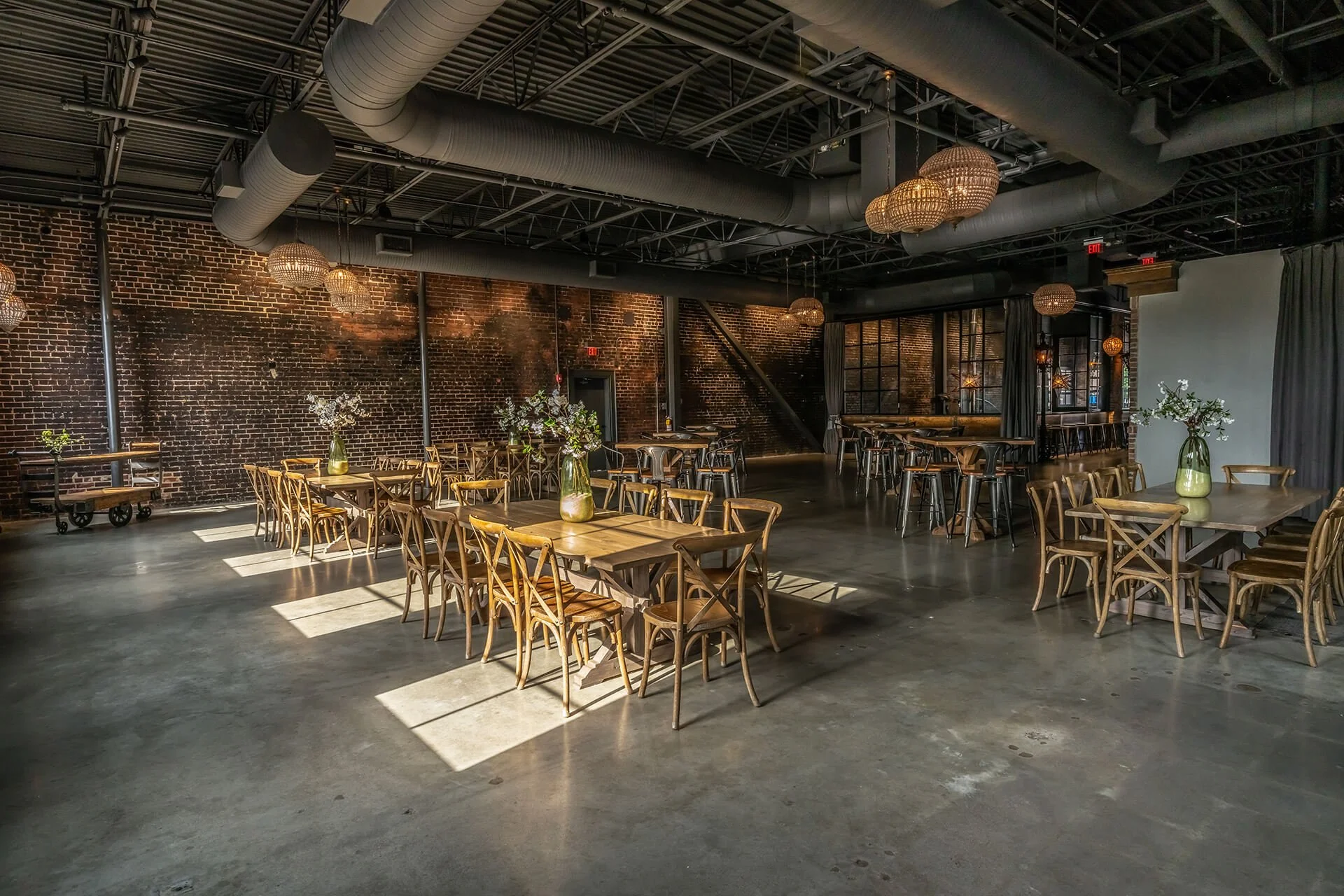 A spacious, modern dining area with wooden tables, chairs, and floral centerpieces under warm lighting against exposed brick walls.