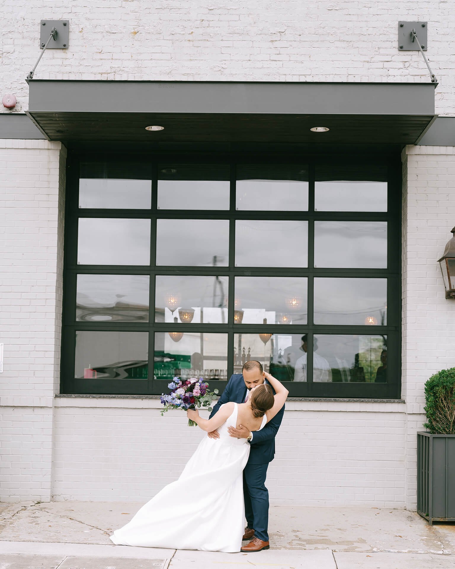 A couple embraces outdoors, with the bride holding a vibrant purple bouquet and wearing a flowing white gown, against an industrial backdrop.