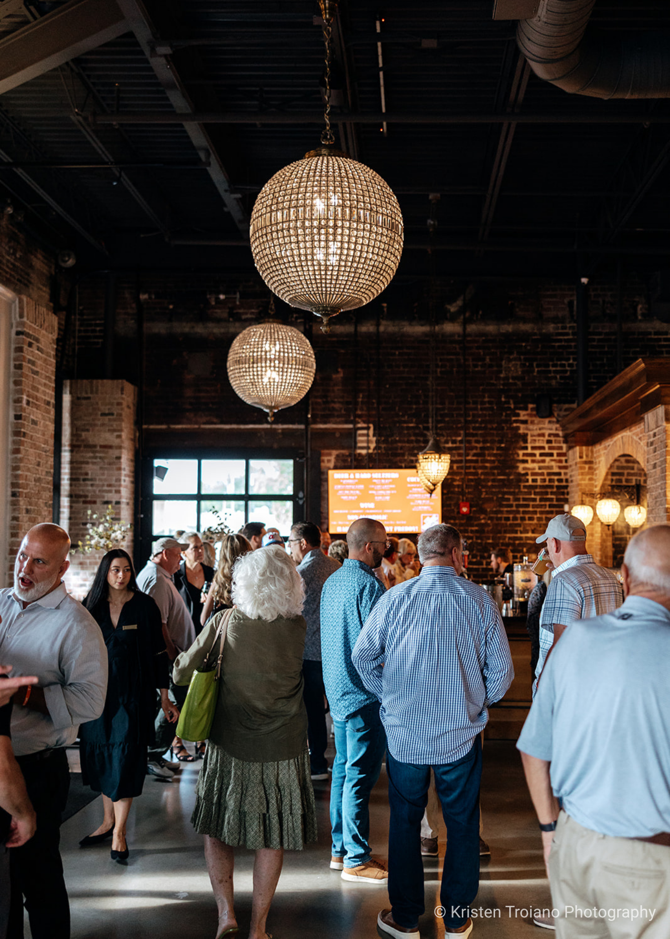 People mingling at an indoor party under crystal chandeliers with a brick wall background.