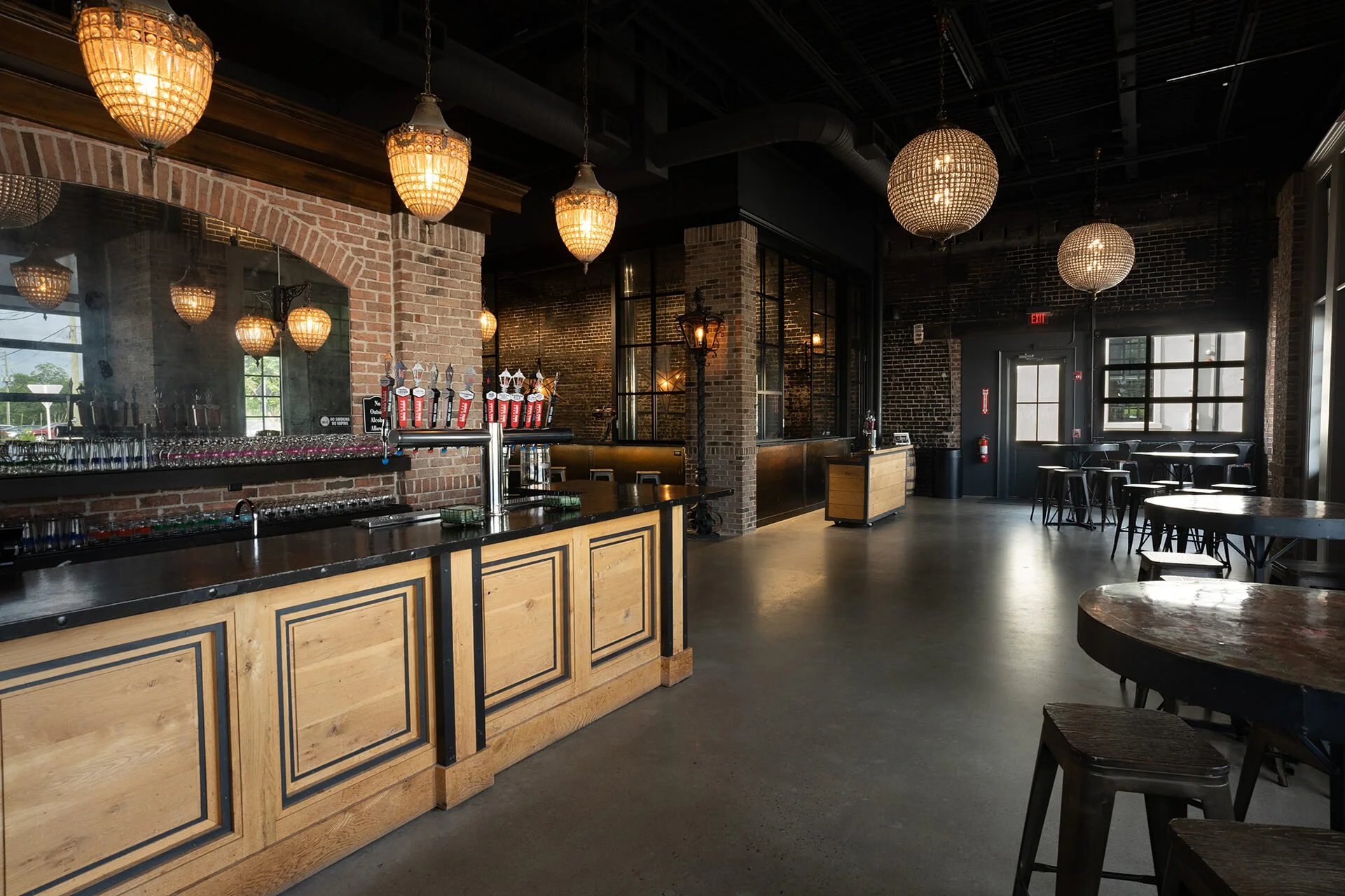 A stylish bar and lounge area with a brick wall backdrop, chandeliers, a sleek counter, and wooden tables in a spacious setting.
