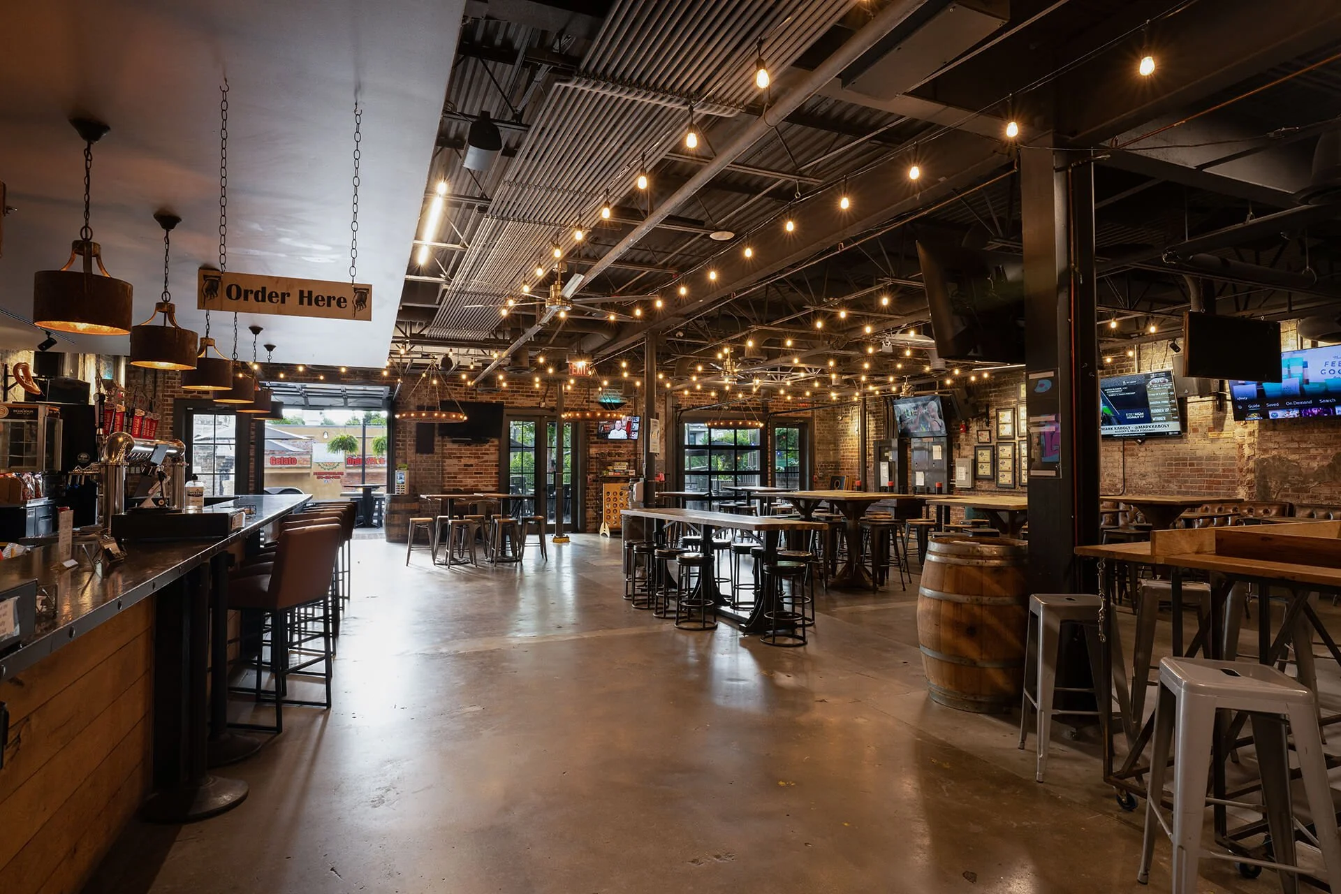  Interior view of a brewery with an industrial design featuring exposed brick walls, a black metal ceiling interspersed with string lights, and a polished concrete floor. The foreground includes a bar area with pendant lights, an "Order Here" sign, a