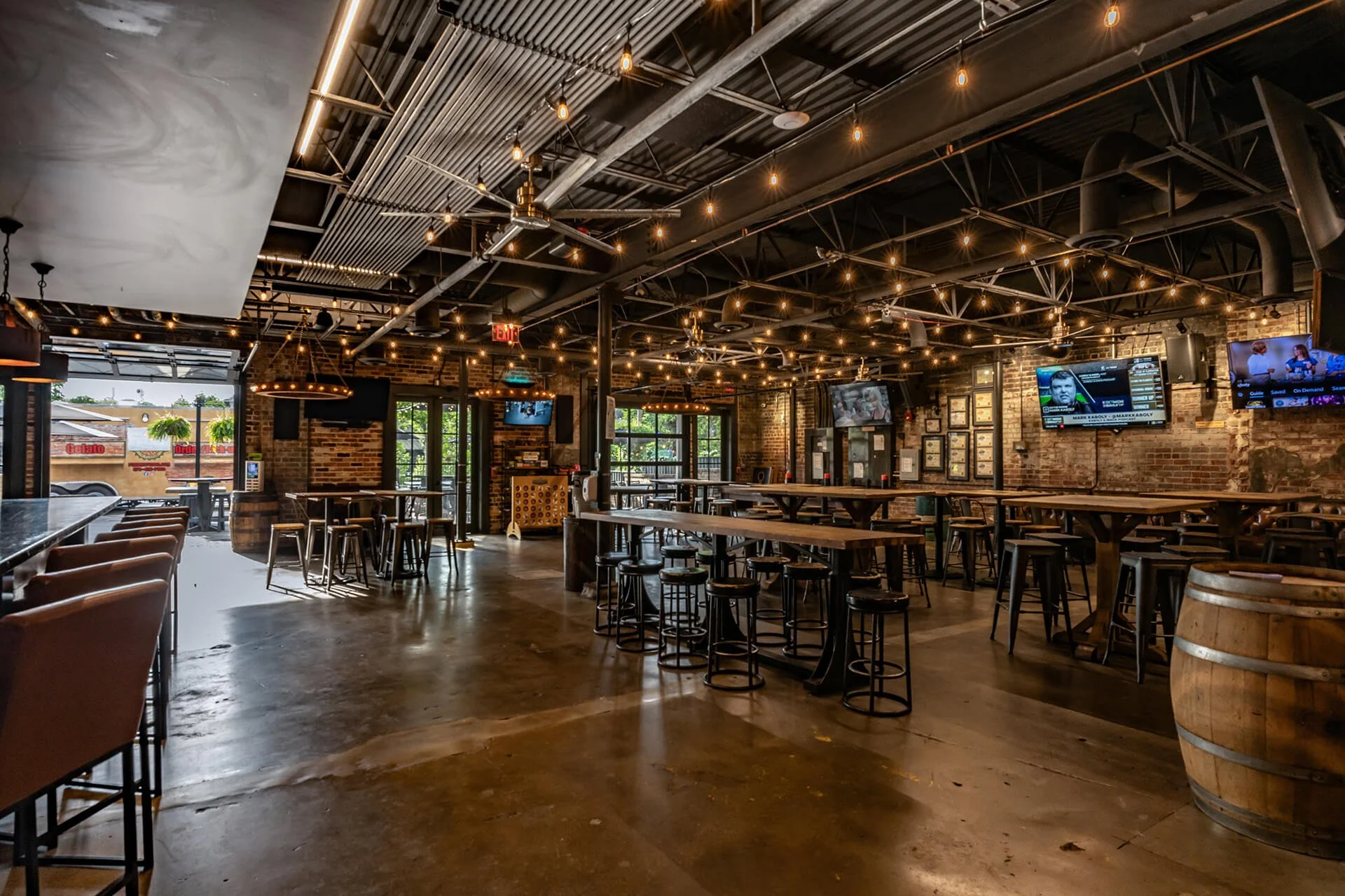  Spacious brewery interior featuring exposed brick walls adorned with multiple televisions, industrial-style lighting strung across a corrugated metal ceiling, and a polished concrete floor reflecting ambient light. Seating arrangements include a lon