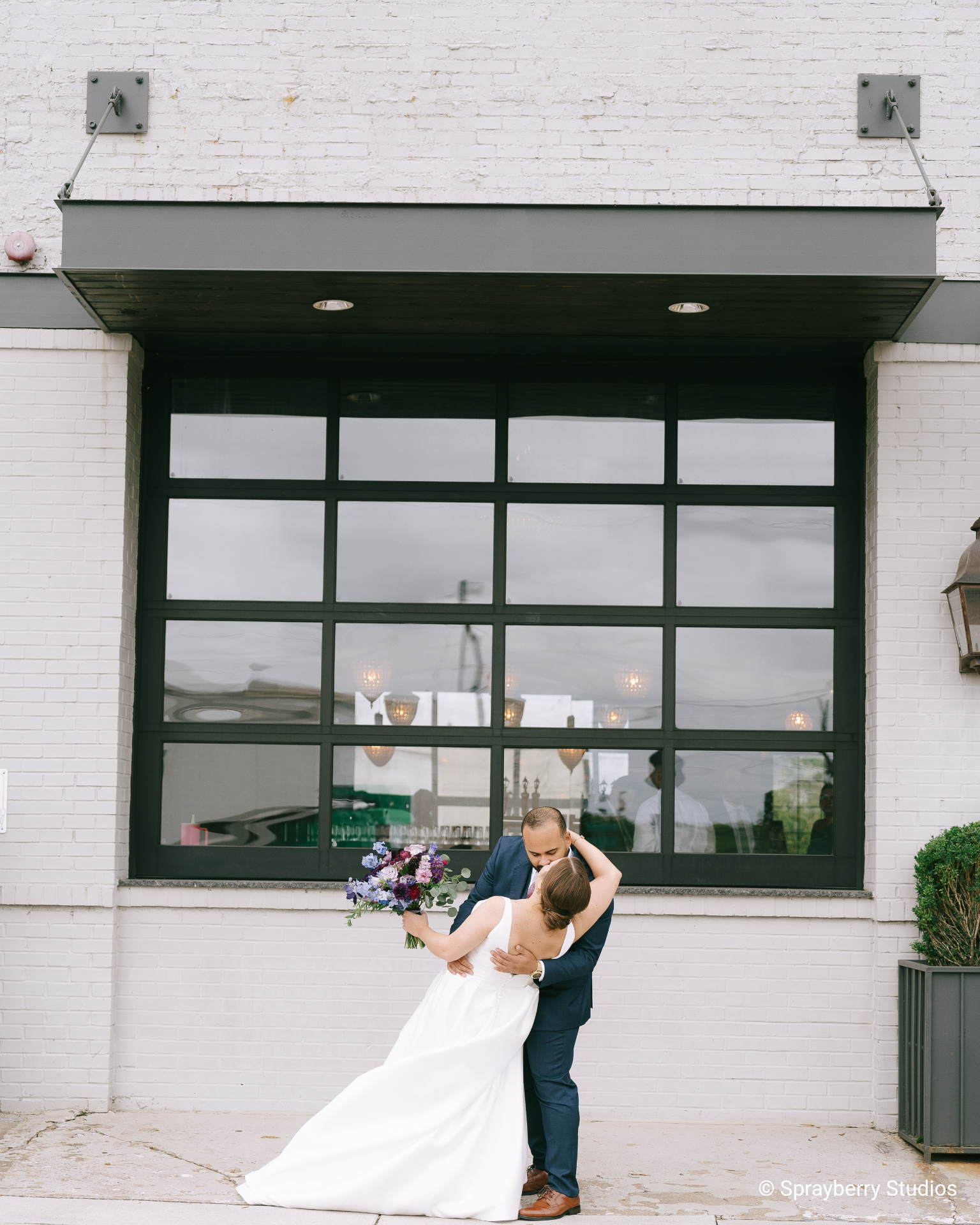 A couple embraces outdoors, with the bride holding a vibrant purple bouquet and wearing a flowing white gown, against an industrial backdrop.
