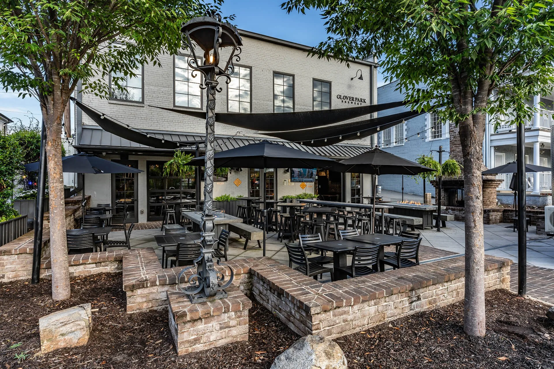  Courtyard seating area for Glover Park Brewery features black tables and chairs arranged under black umbrellas and a stretched shade awning attached to brewery building. Light-colored brick building has matching light-colored trim around dark window