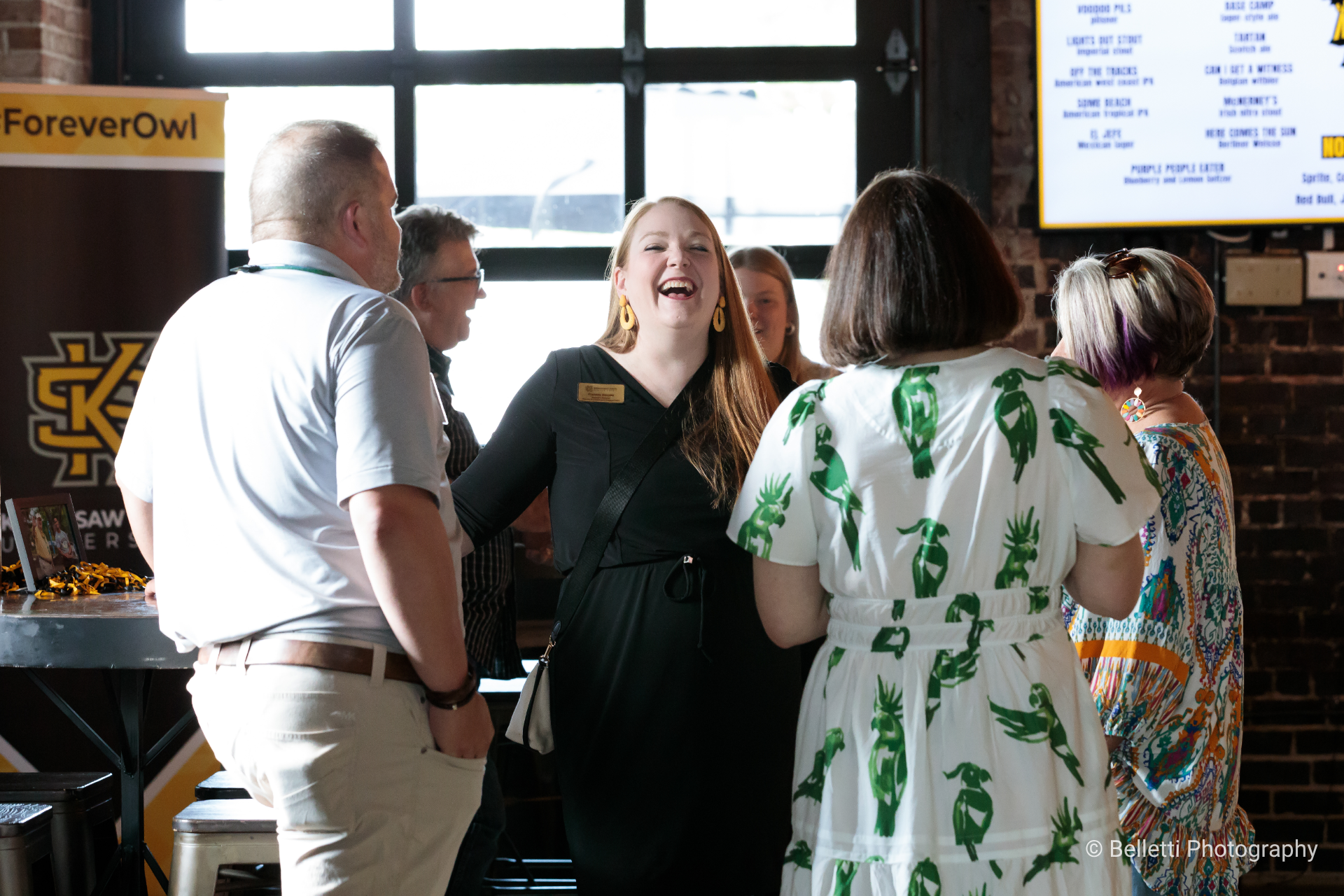 Folks gather and chat at an event; a woman in a black dress and bright yellow earrings laughs openly.