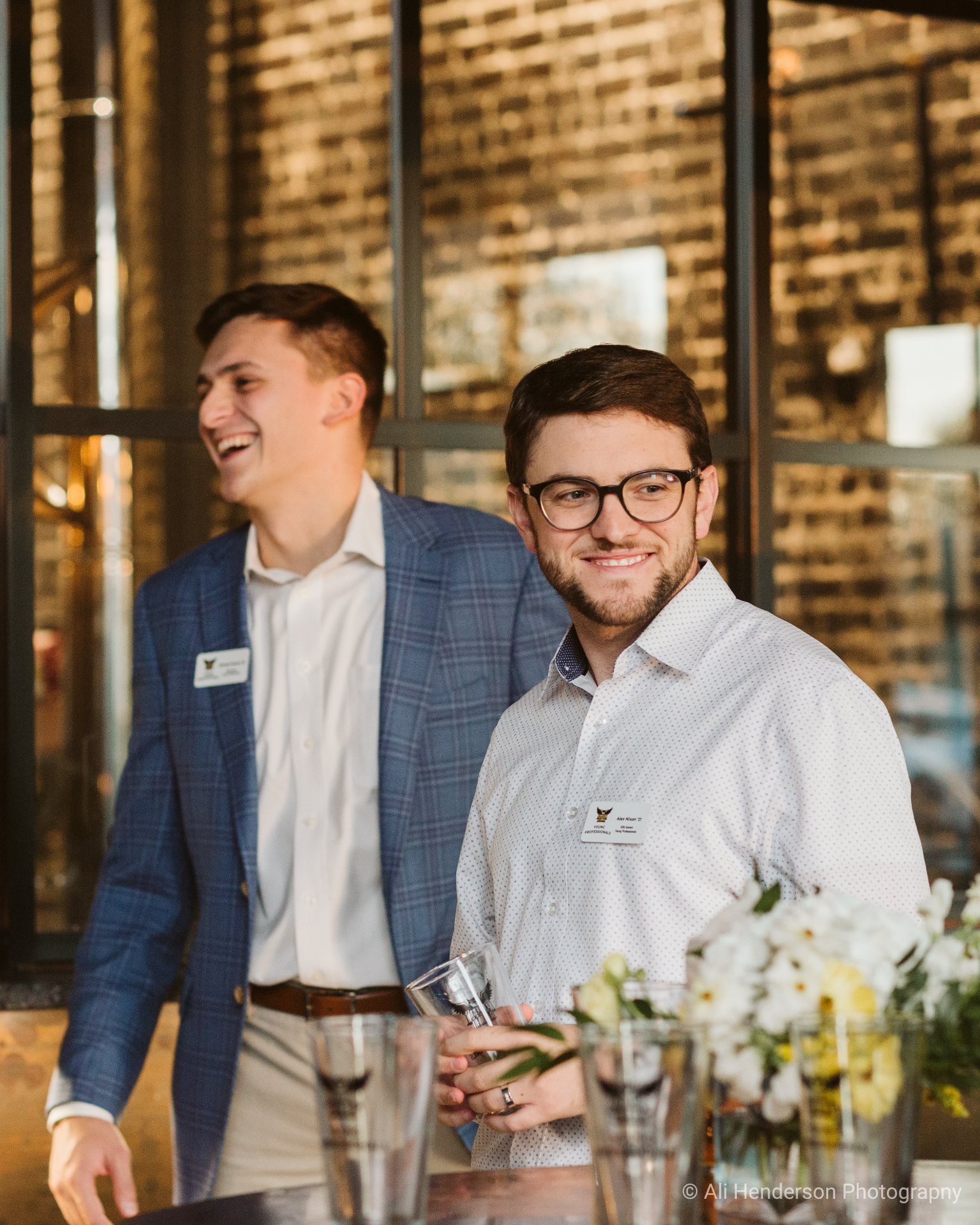 Two men stand at a bar, one in a blue plaid jacket and the other in a white button-up shirt, surrounded by empty glasses and flowers.