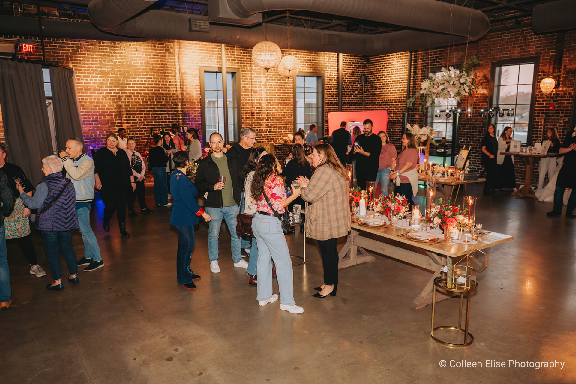 Party scene in loft-like venue with exposed brick walls, concrete floors, and hanging lights, with diverse crowd of people chatting and enjoying themselves, some around a long wooden banquet table adorned with florals and candles.