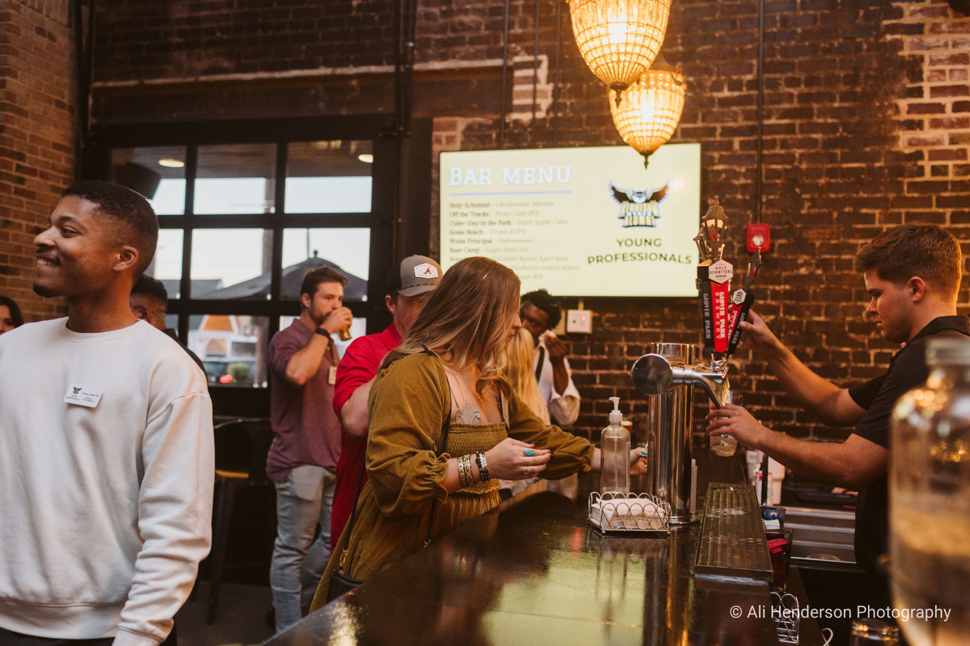 At a crowded party, a woman in an olive dress fills her glass at the bar while smiling. A bar menu is visible on the wall, and exposed brick adds to the ambiance.