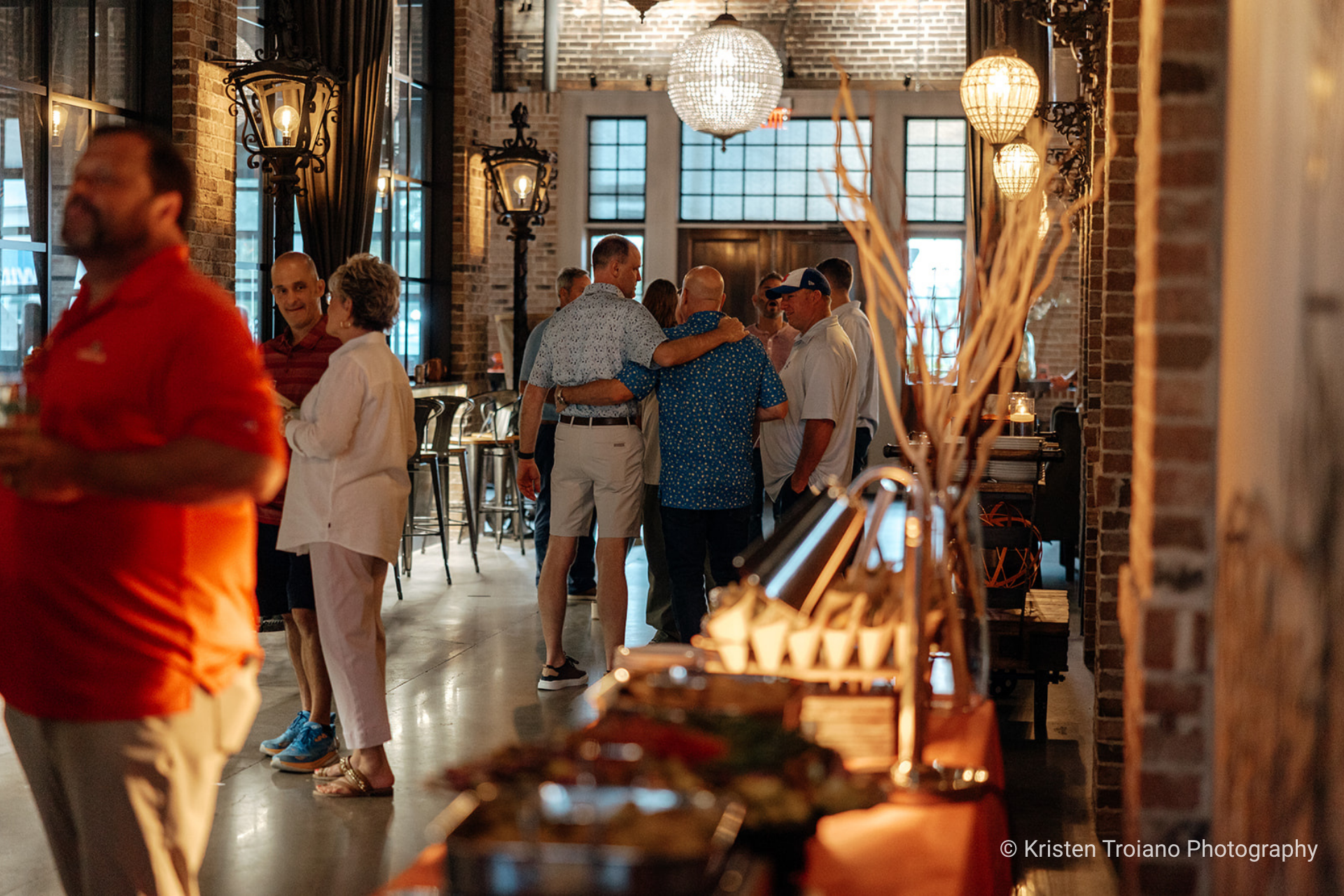 People gathered at a party inside a brick building. Some are chatting, and food is displayed in the foreground.