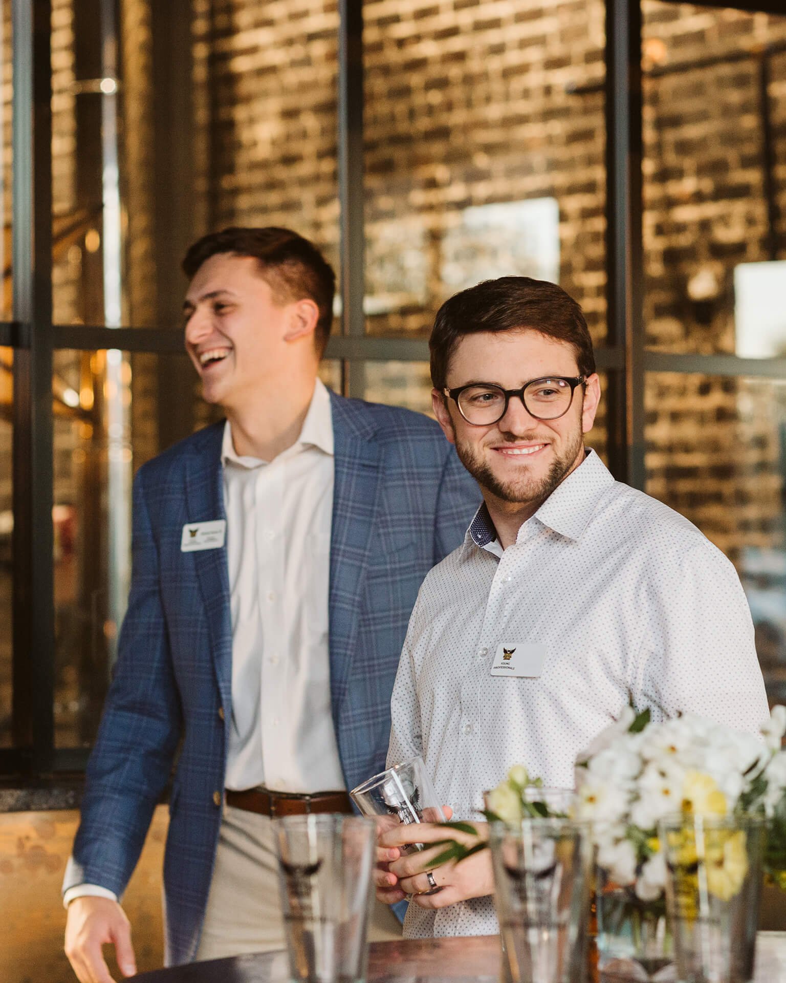Two men stand at a bar, one in a blue plaid jacket and the other in a white button-up shirt, surrounded by empty glasses and flowers.