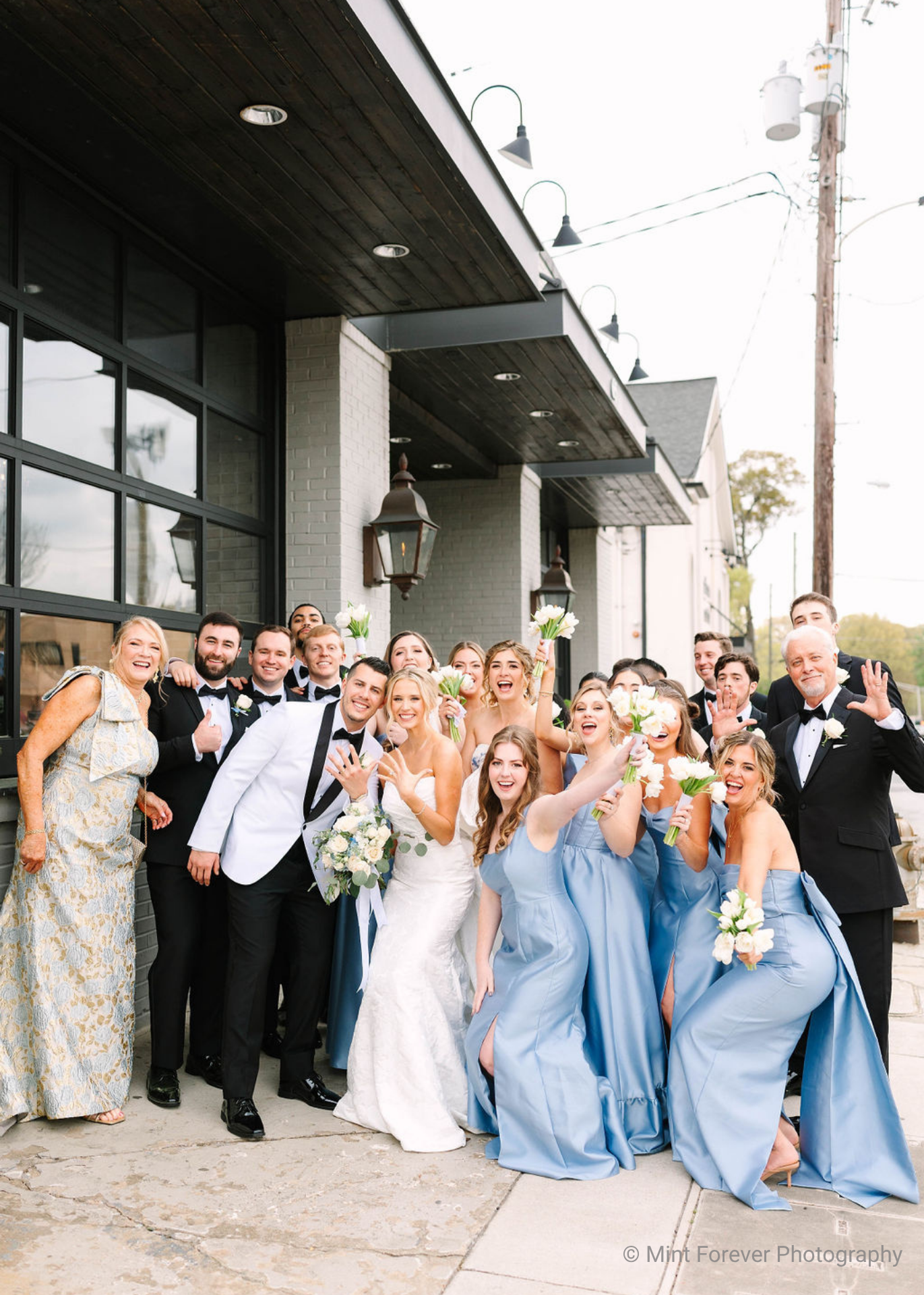 Wedding party posing outdoors; bride and groom displaying wedding rings, surrounded by bridesmaids in light blue gowns and groomsmen in black tuxedos, all smiling and cheerful.