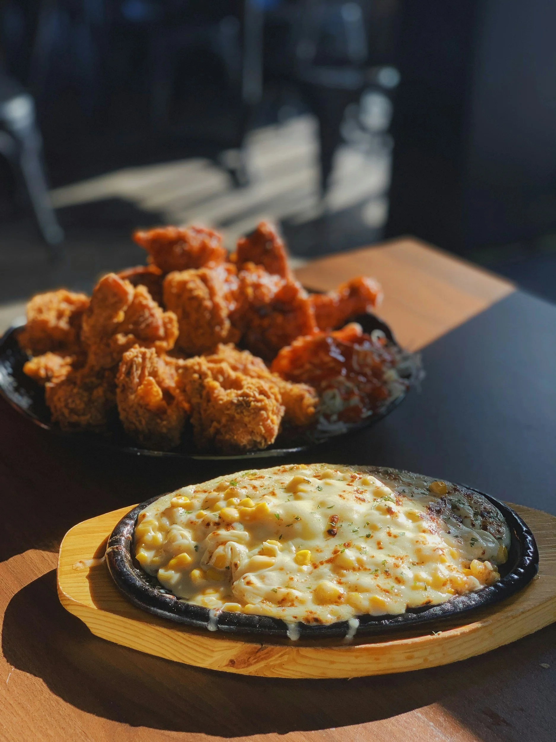 A sizzling plate of cheesy corn casserole and a black plate of fried chicken with various sauces on a wooden table.