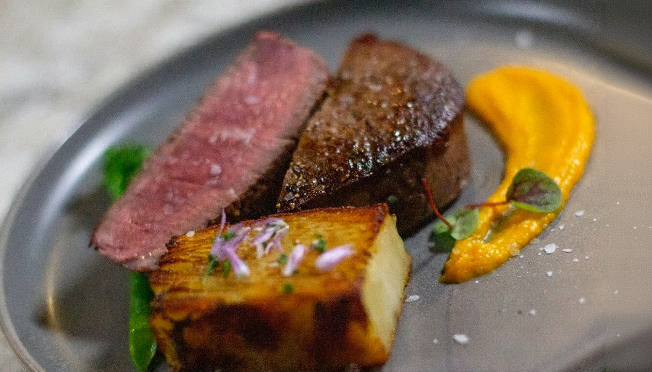 A plated dish with a slice of pink steak, a piece of seared meat, roasted potato with herbs, and a smear of yellow sauce garnished with a small green herb and pink flower petals.
