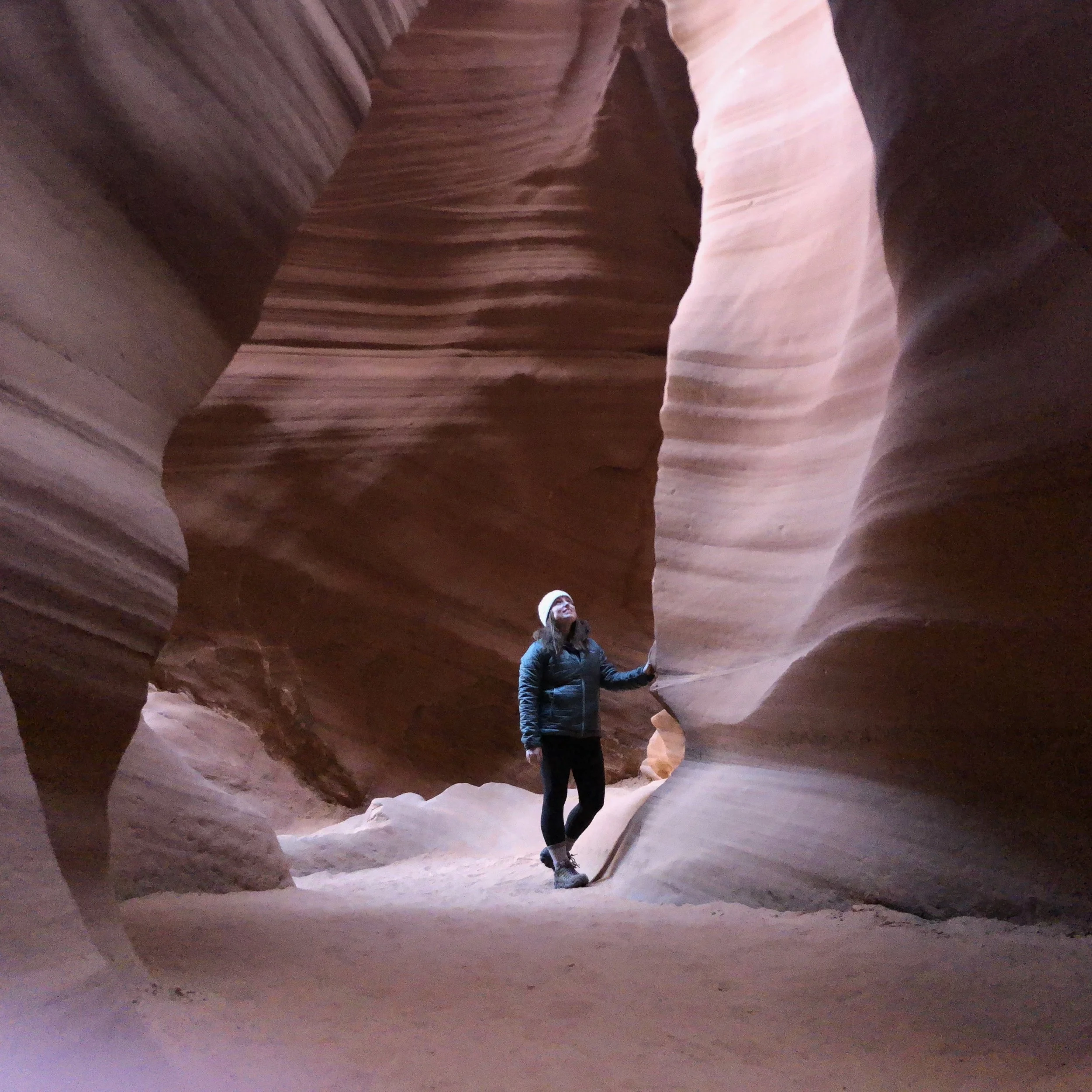 A person wearing a white hat, black jacket, and pants standing inside a narrow slot canyon with smooth, layered sandstone walls.