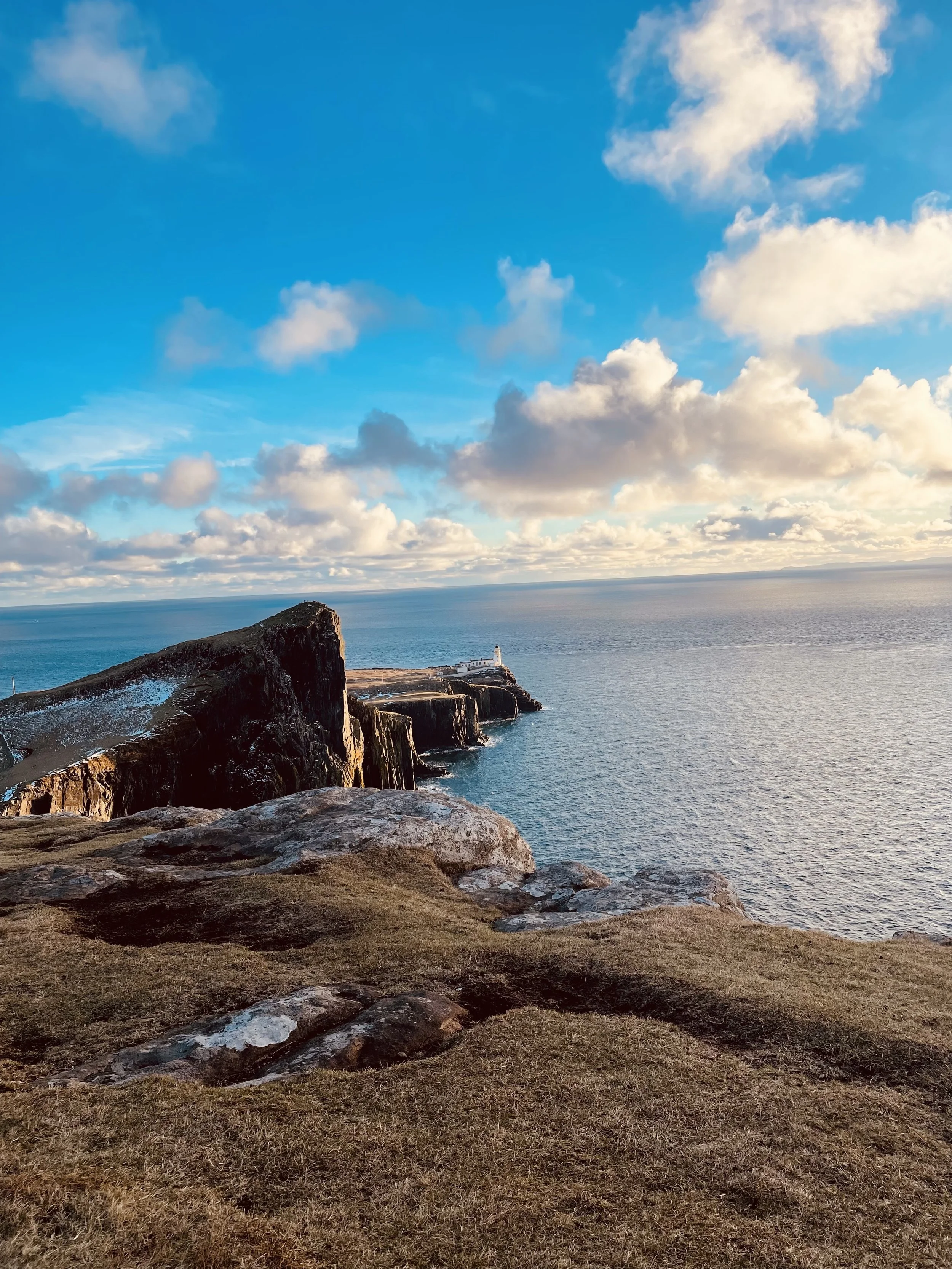 Scenic view of the rugged coastline of Neist Points on the Isle of Skye in Scotland with cliffs and the ocean, featuring a lighthouse at the tip of the landmass, under a partly cloudy sky with sunlight illuminating the scene.