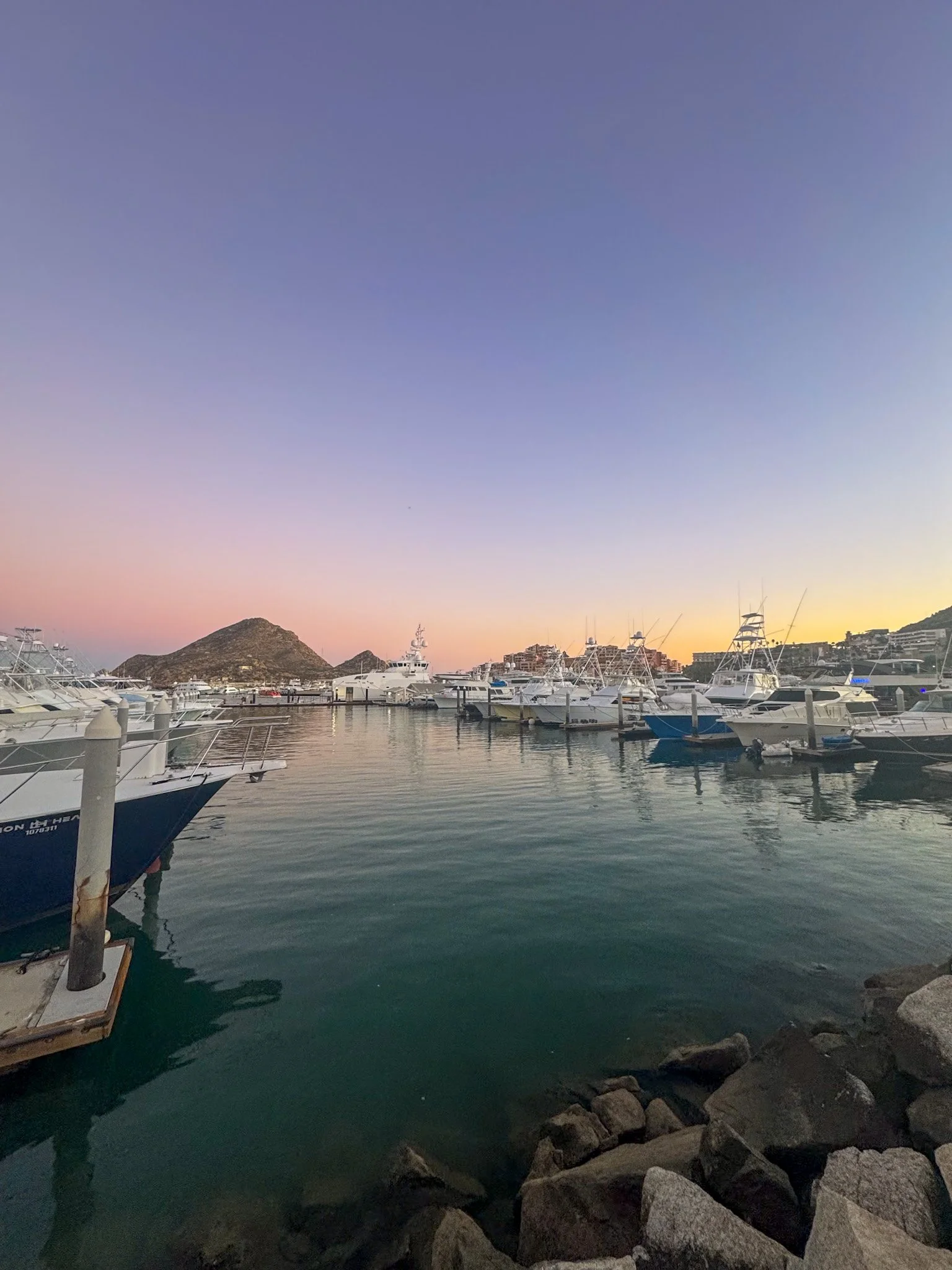 a pink and purple sunset over the cabo san lucas marina with mount solar in the background