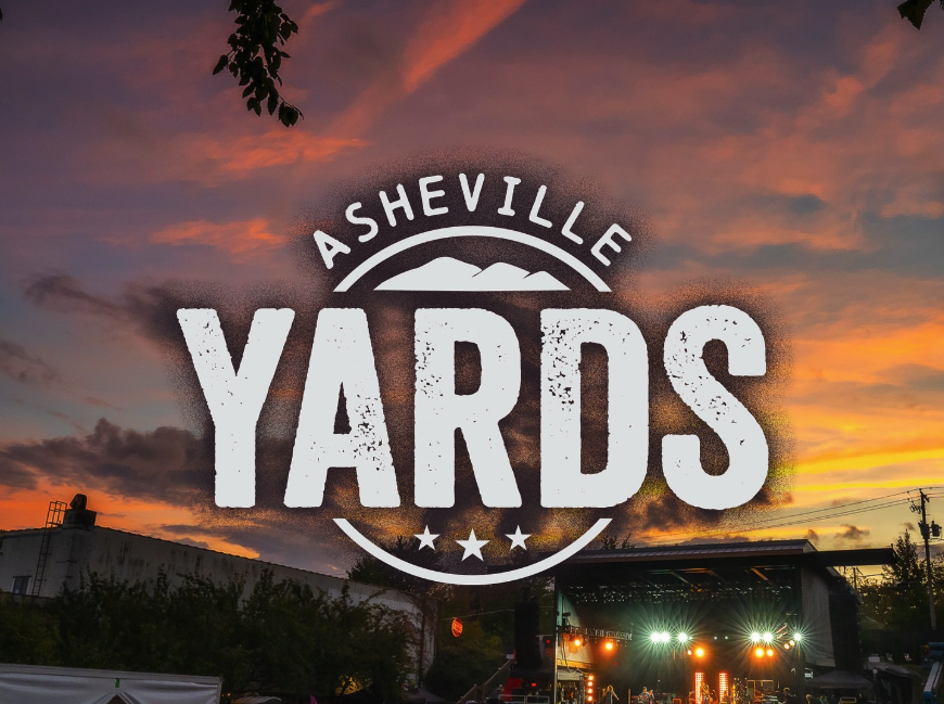 Sunset sky with orange and purple clouds over an outdoor concert stage, with buildings, trees, and power lines visible in the foreground, and a logo reading 'Asheville Yards' centered in the image.