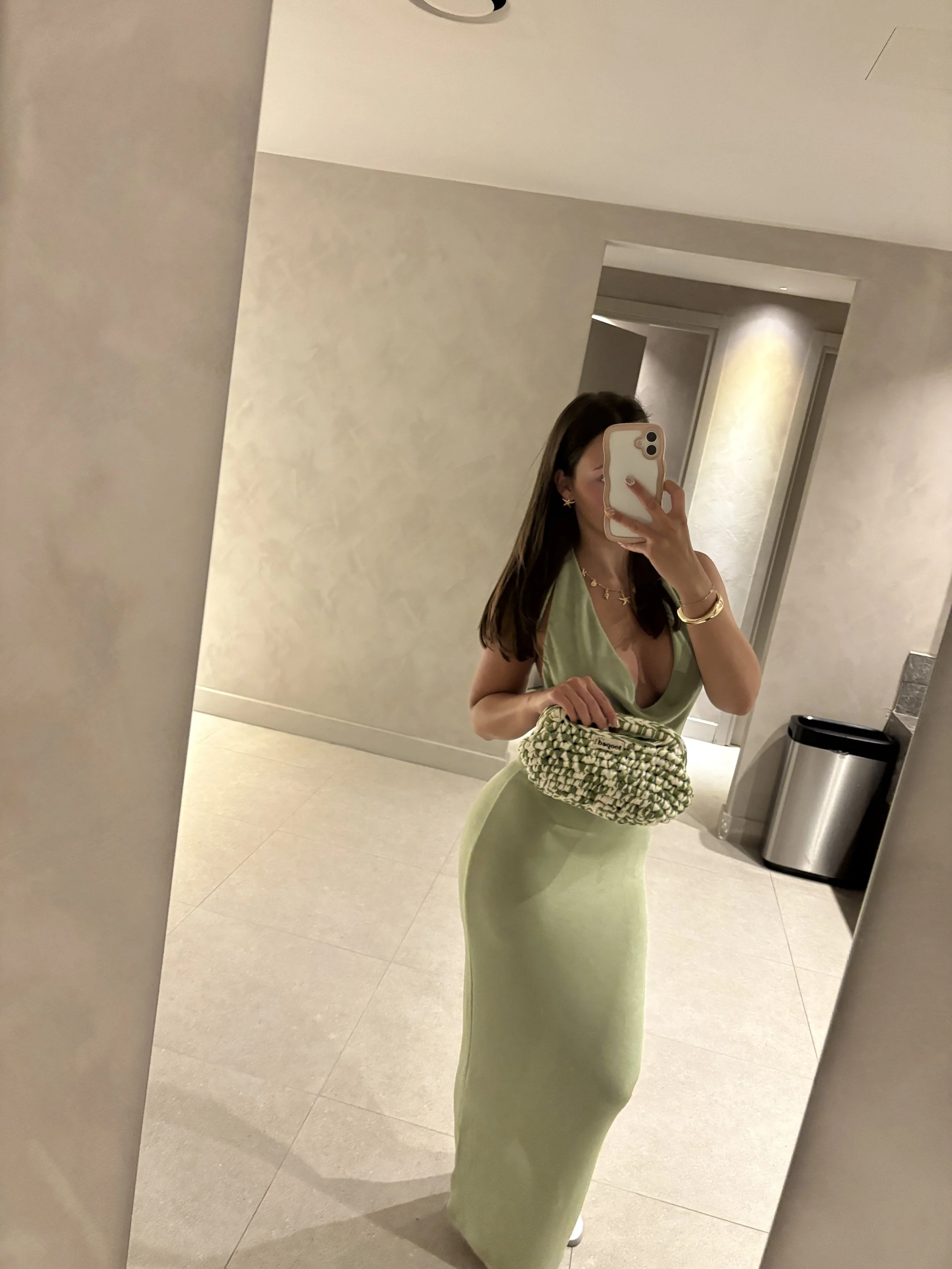 Woman wearing a light green dress taking a mirror selfie in a modern indoor setting, holding a patterned handbag, with a trash can visible in the background.