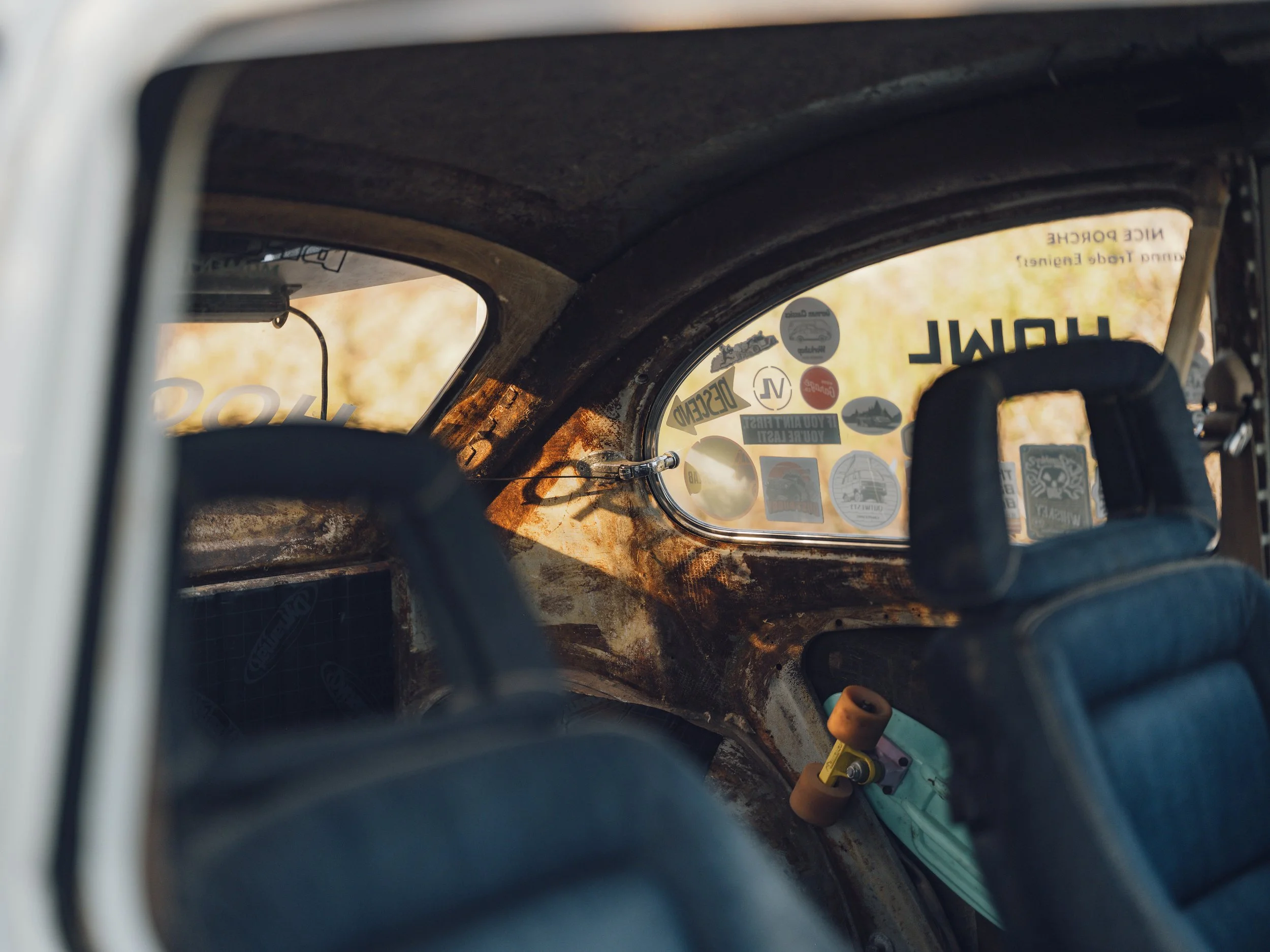 The interior of a vintage car showing two black racing seats, a rusty dashboard, a small windshield with various stickers, and a mini skateboard with blue deck and orange wheels.