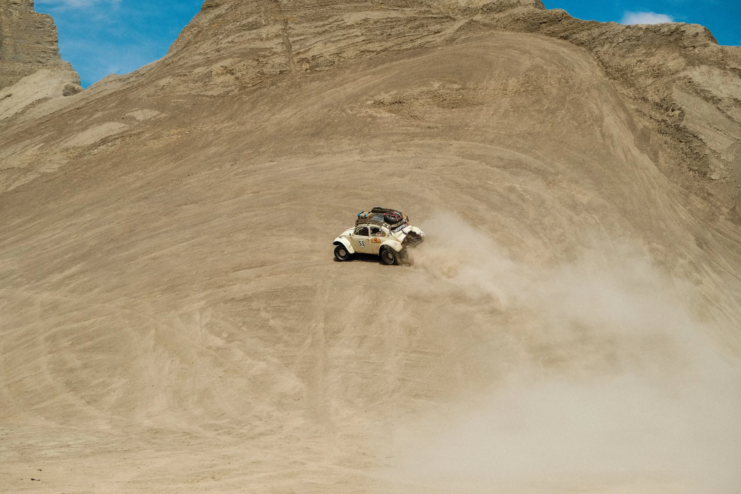 A vintage off-road vehicle racing on a steep, sandy desert slope, kicking up dust with a mountainous background under a blue sky.