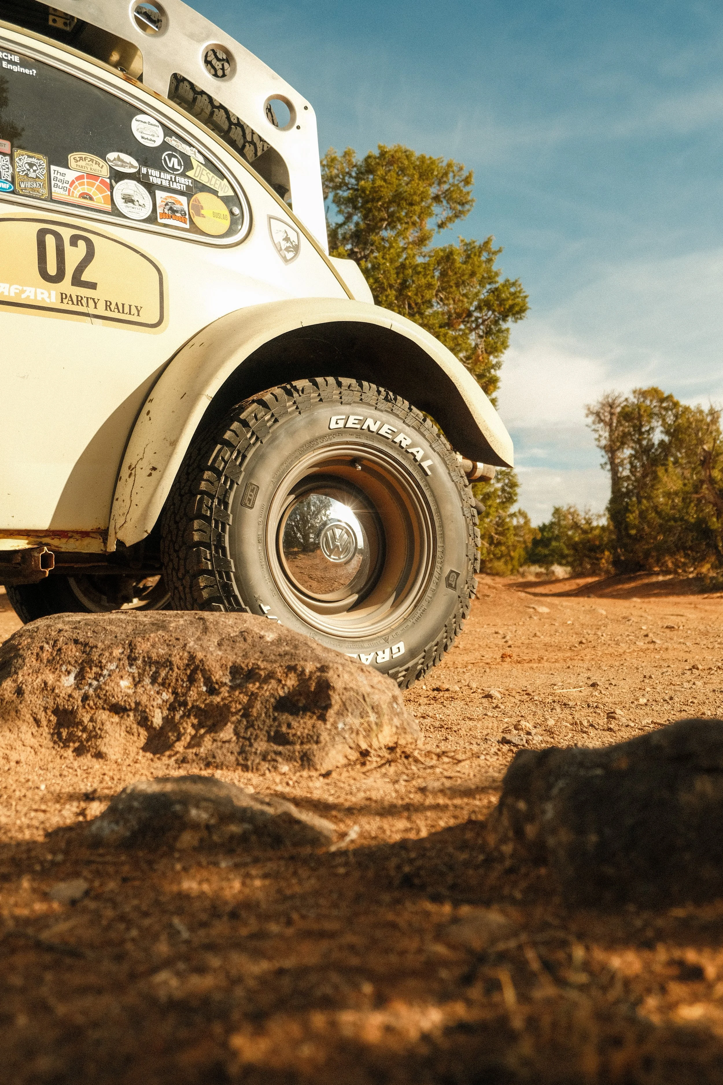 Close-up of a vintage white Volkswagen vehicle on a dirt trail with stickers on the windshield, rocks in the foreground, and trees in the background under a clear blue sky.