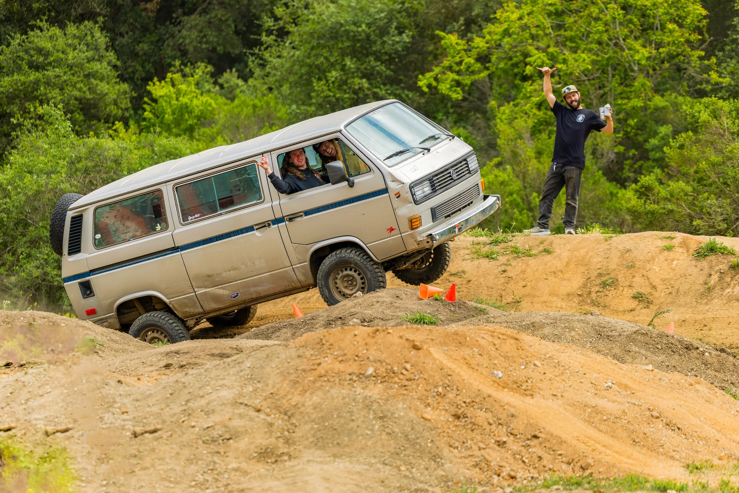 A beige Volkswagen van off-roading on a dirt track with two people inside smiling and waving, while a man standing nearby holding a drink and raising a hand in celebration, surrounded by green trees.