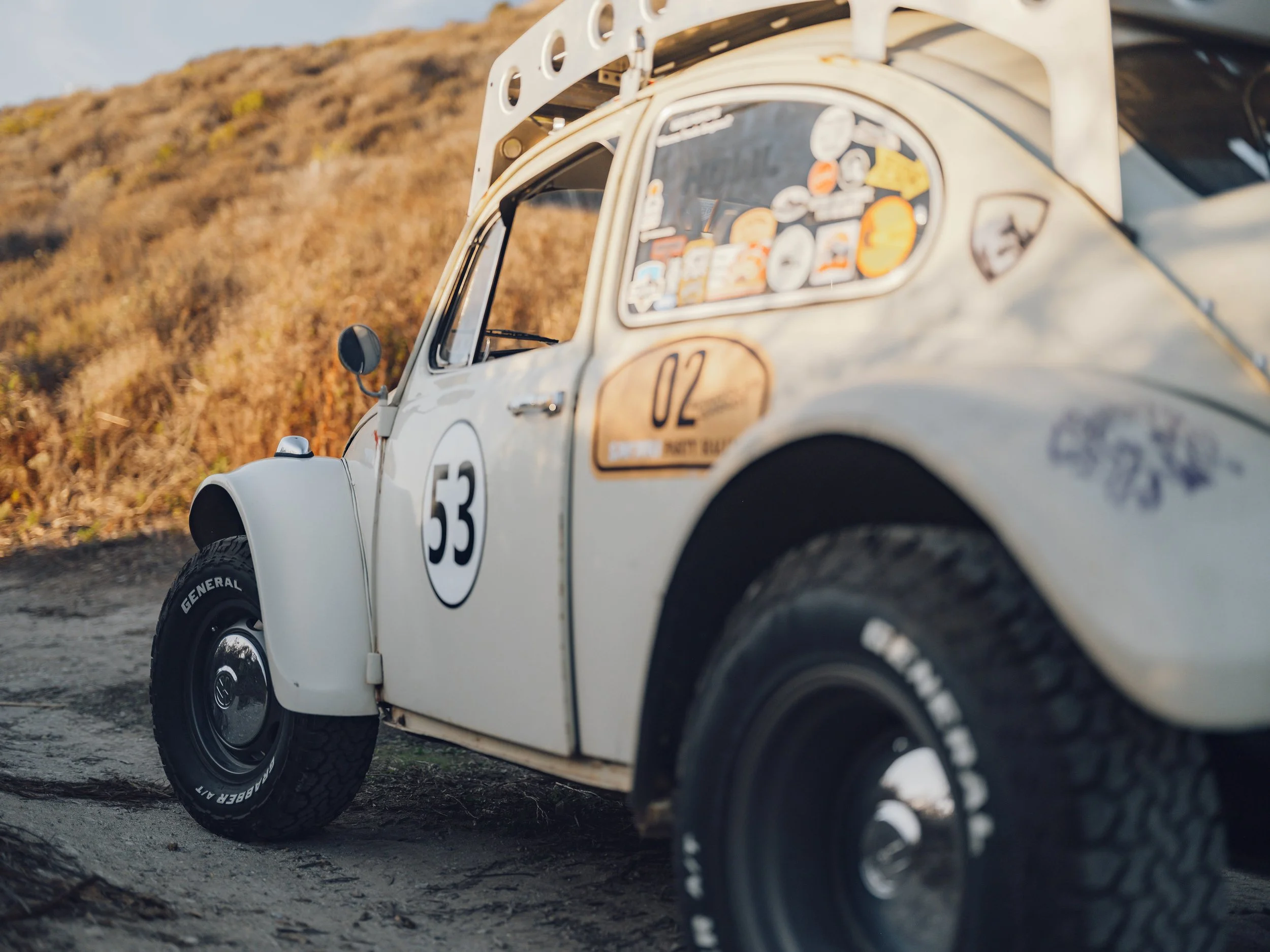 A vintage white Volkswagen Beetle rally car with the number 53 painted on the side, parked on a dirt road with a hillside in the background during sunset.
