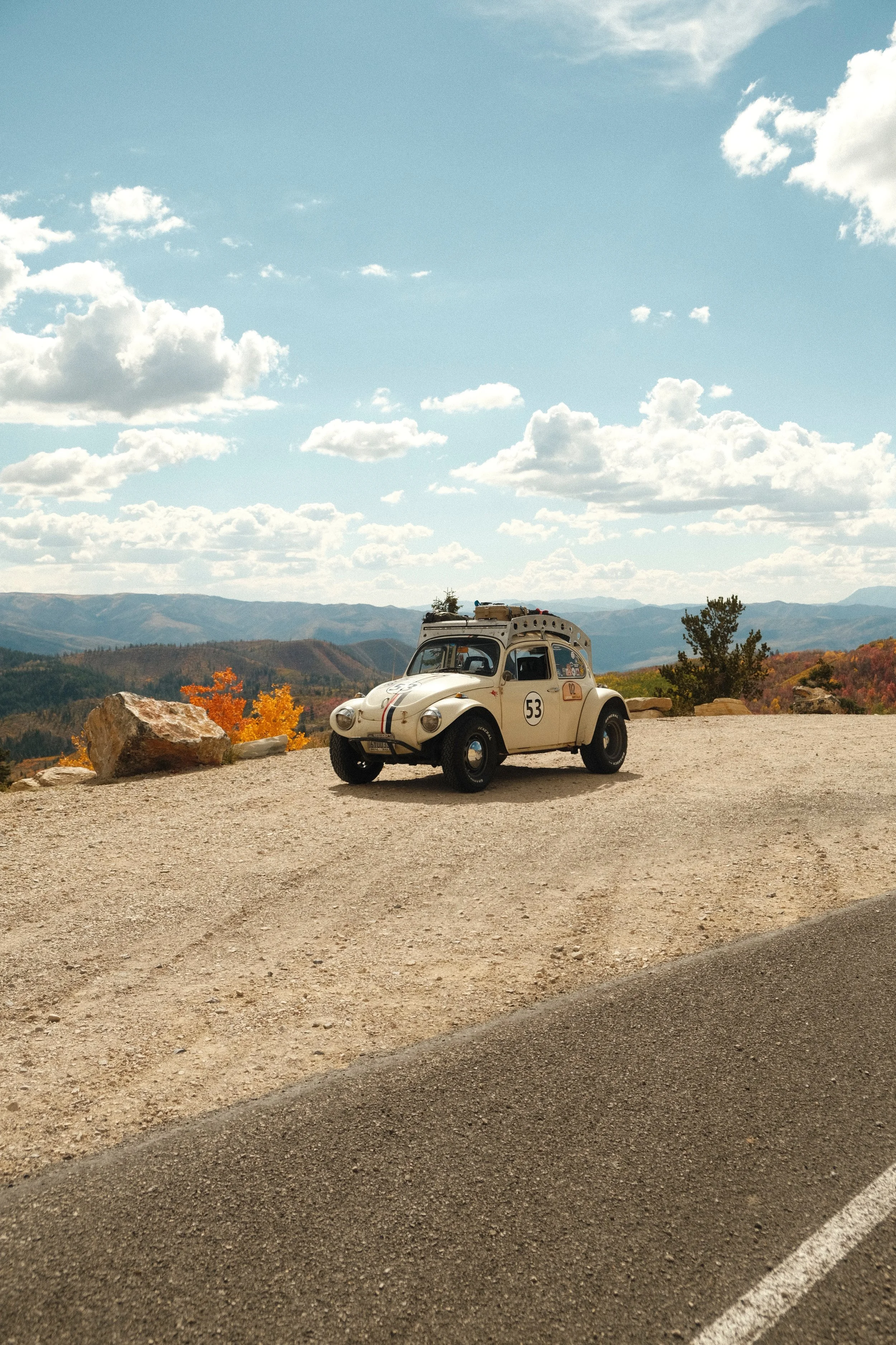 A vintage white Volkswagen Beetle with racing stripes and racing decals parked on a scenic mountain overlook during daytime with partly cloudy sky and rolling hills in the background.