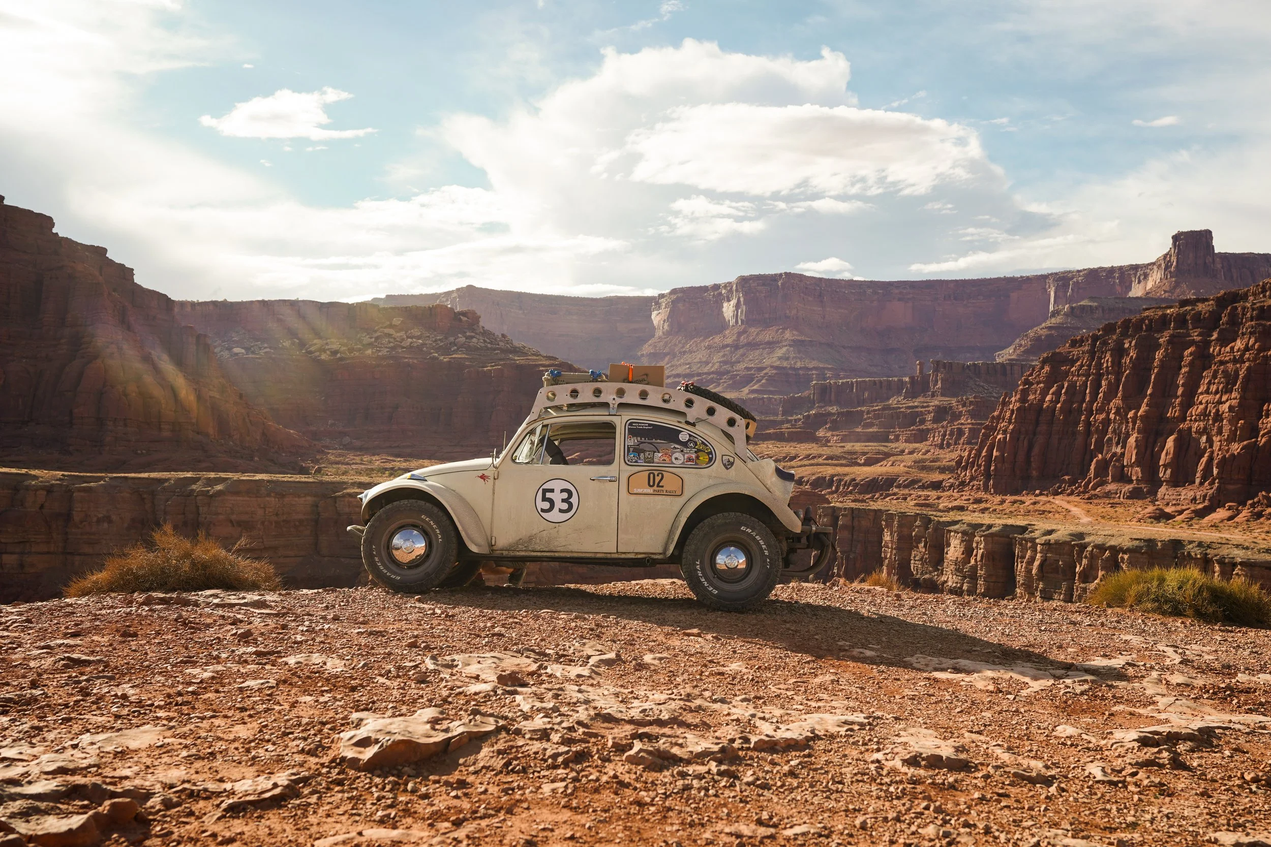 A vintage toy polar beige car with racing stickers and the number 53 on its door, parked on a rocky outcrop overlooking a canyon landscape, with a partly cloudy sky overhead.