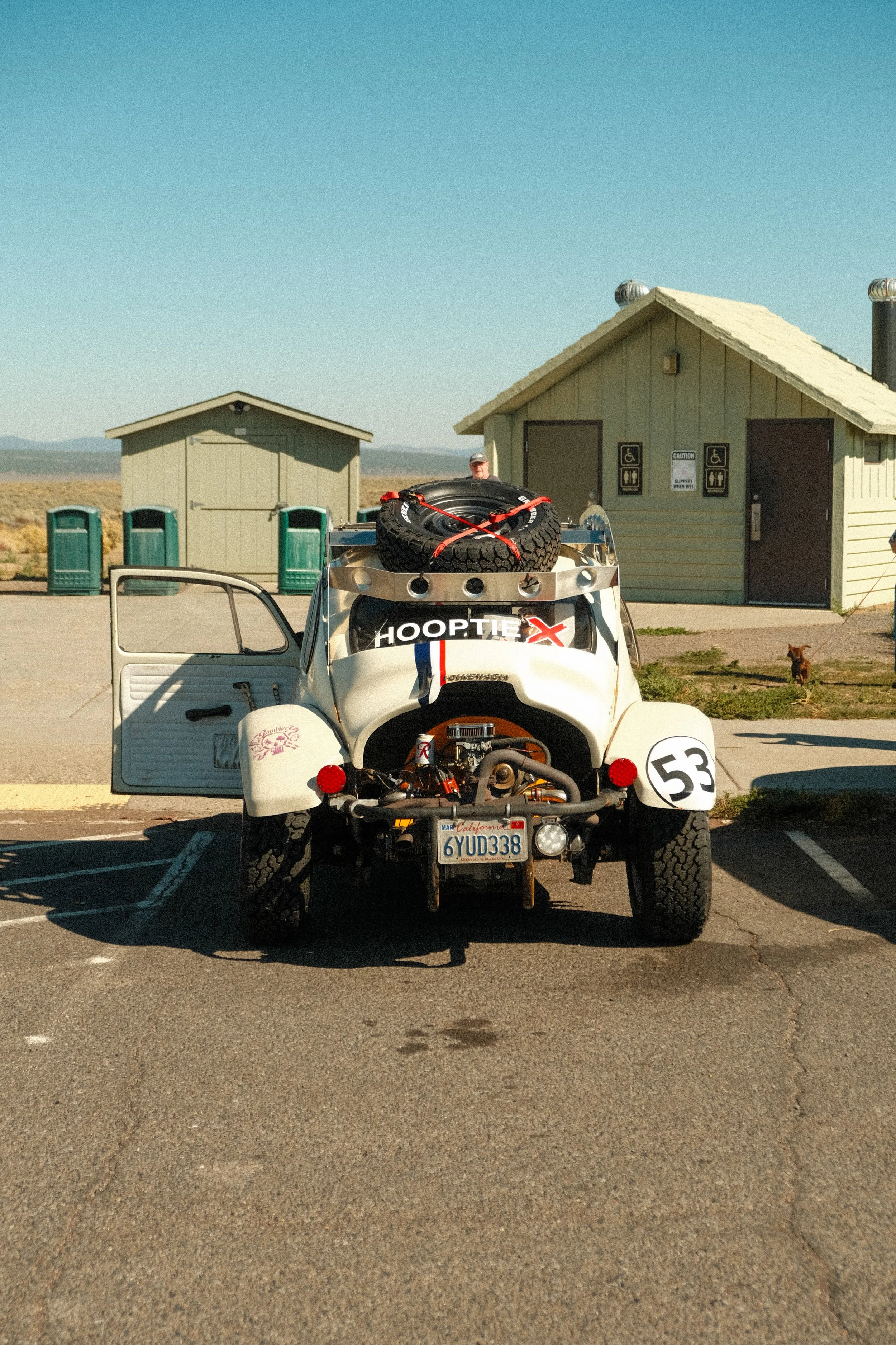 A vintage dune buggy with a spare tire on top and the number 53 on the right fender, parked in a lot near a small building with signs for handicapped accessibility. The car has its driver's side door open, and the license plate reads '6YUD338'. The b