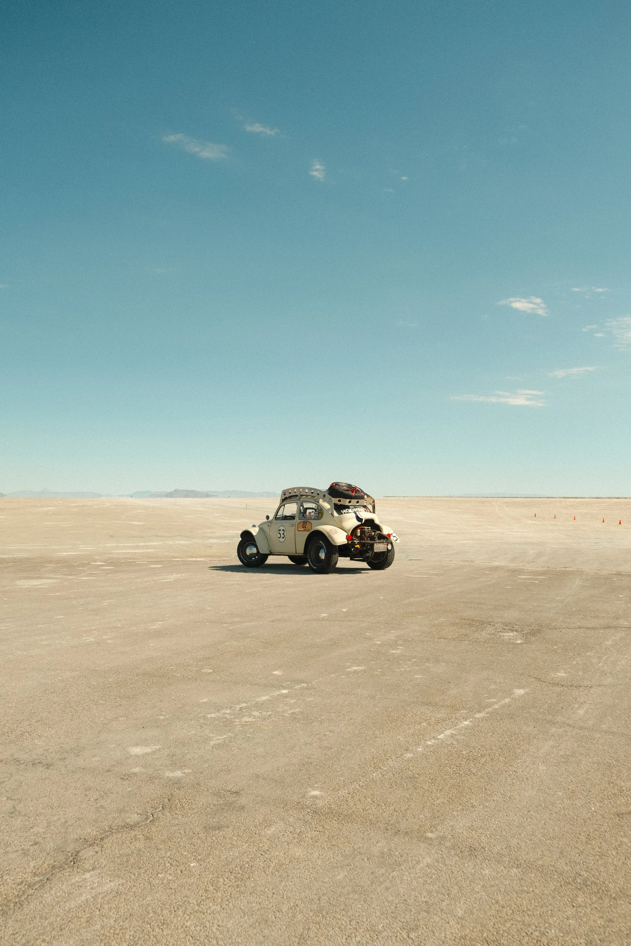 A vintage white car with racing decals and luggage on top is parked alone on a vast, flat salt flats under a clear blue sky.