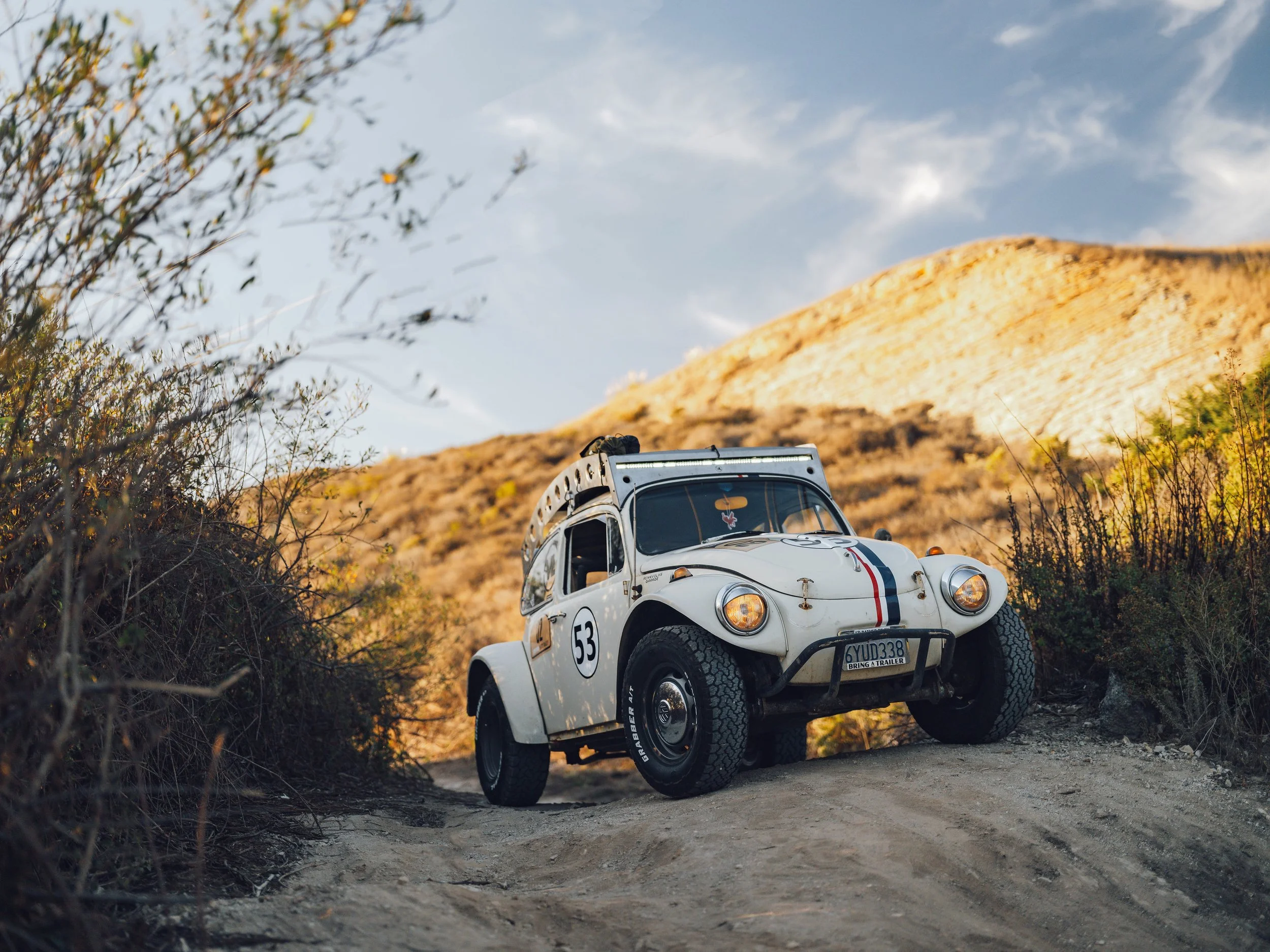 A vintage white off-road vehicle with racing decals and the number 53, driving on a rugged dirt trail through a hilly, desert landscape under a partly cloudy sky.