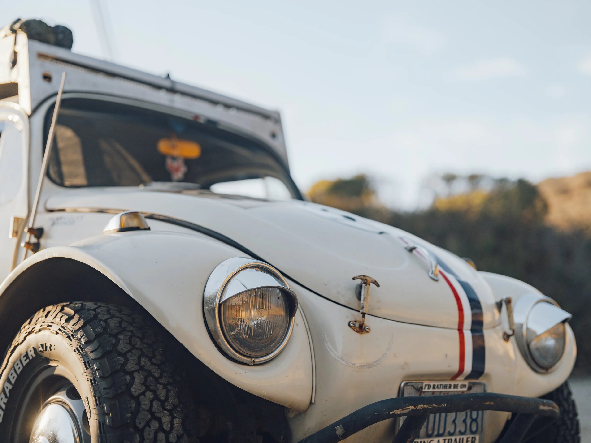 Close-up of a vintage off-road race car with rugged tires, white body with racing stripes, rusted hooks, and a worn license plate, parked outdoors during daylight.