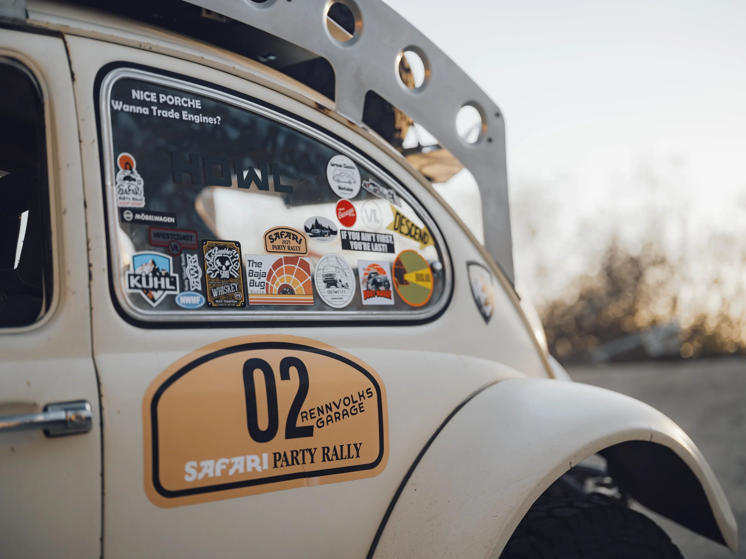Close-up of a vintage beige off-road vehicle with several stickers on the window and body. The window has various travel and rally stickers, including a large sticker that reads '02 RENVOLKS GARAGE SAFARI PARTY RALLY'. The background shows a blurry o