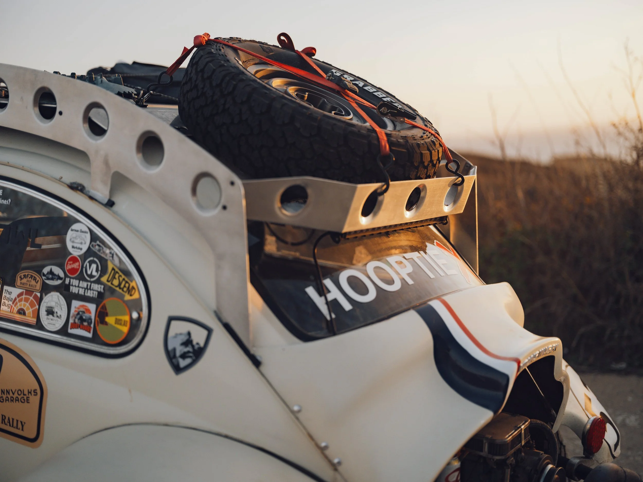 Close-up of a vintage rally car with a spare tire on a metal rack on its roof, adorned with stickers and decals, during sunset.