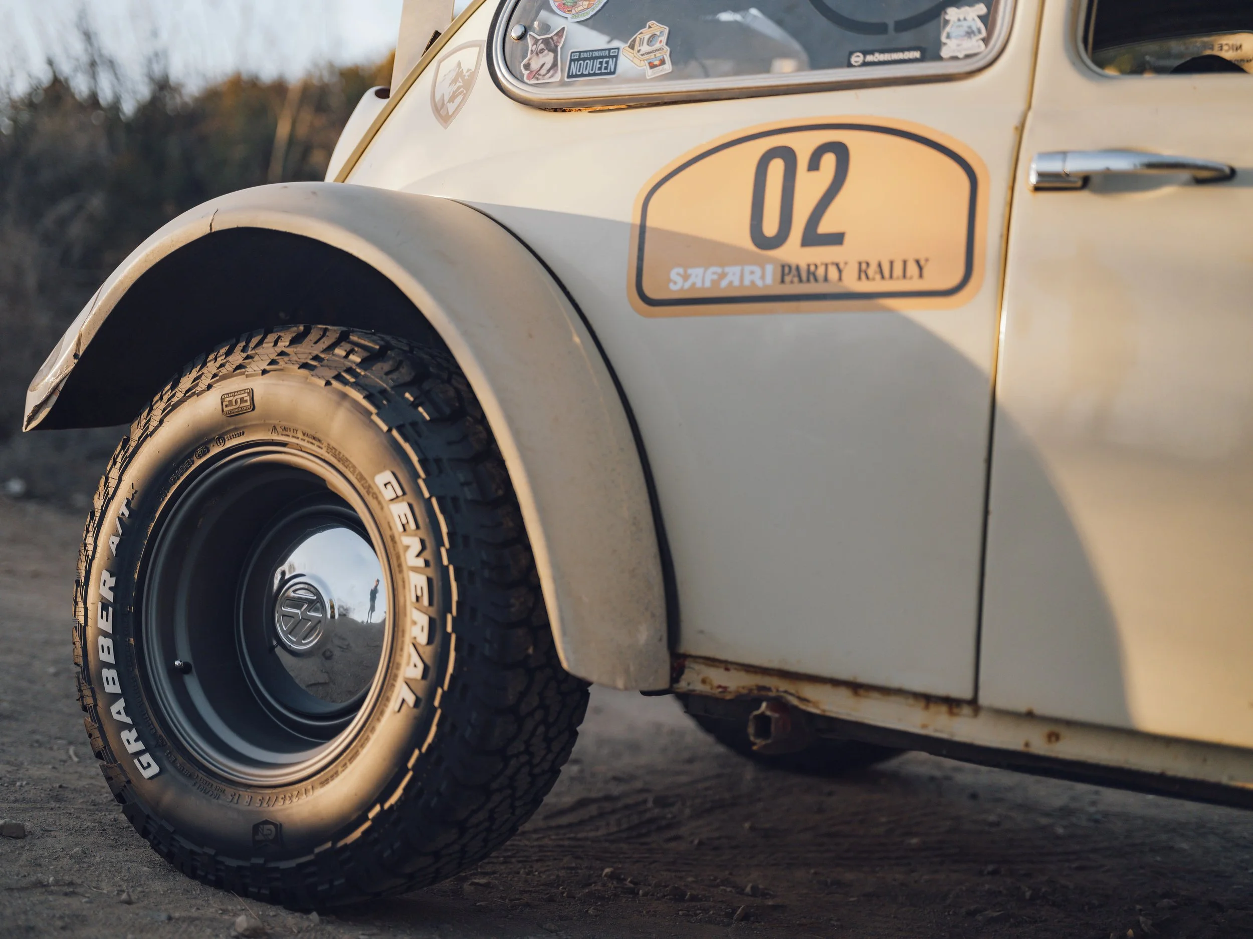 Close-up of the front left side of a vintage beige Volkswagen Beetle, showing its tire with distinctive tread, hubcap, and a rally sticker on the door that reads '02 SAFARI PARTY RALLY'.