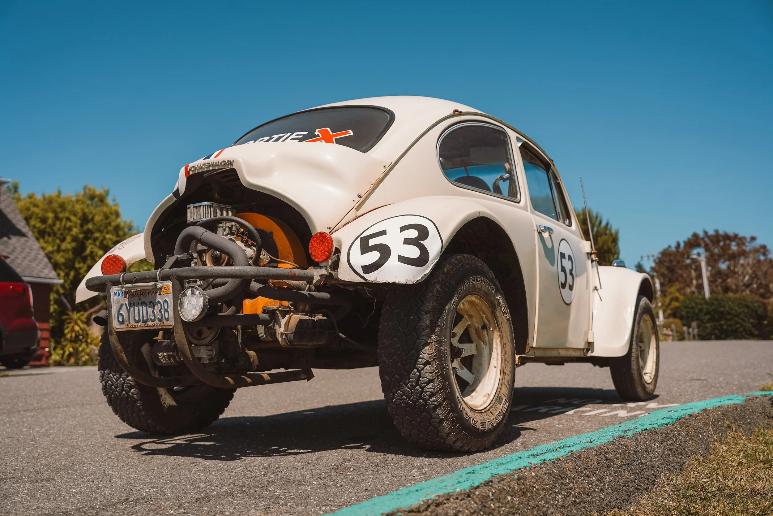 An old white Volkswagen Beetle car with the number 53 on the door and hood, parked on an asphalt street with a blue sky background.
