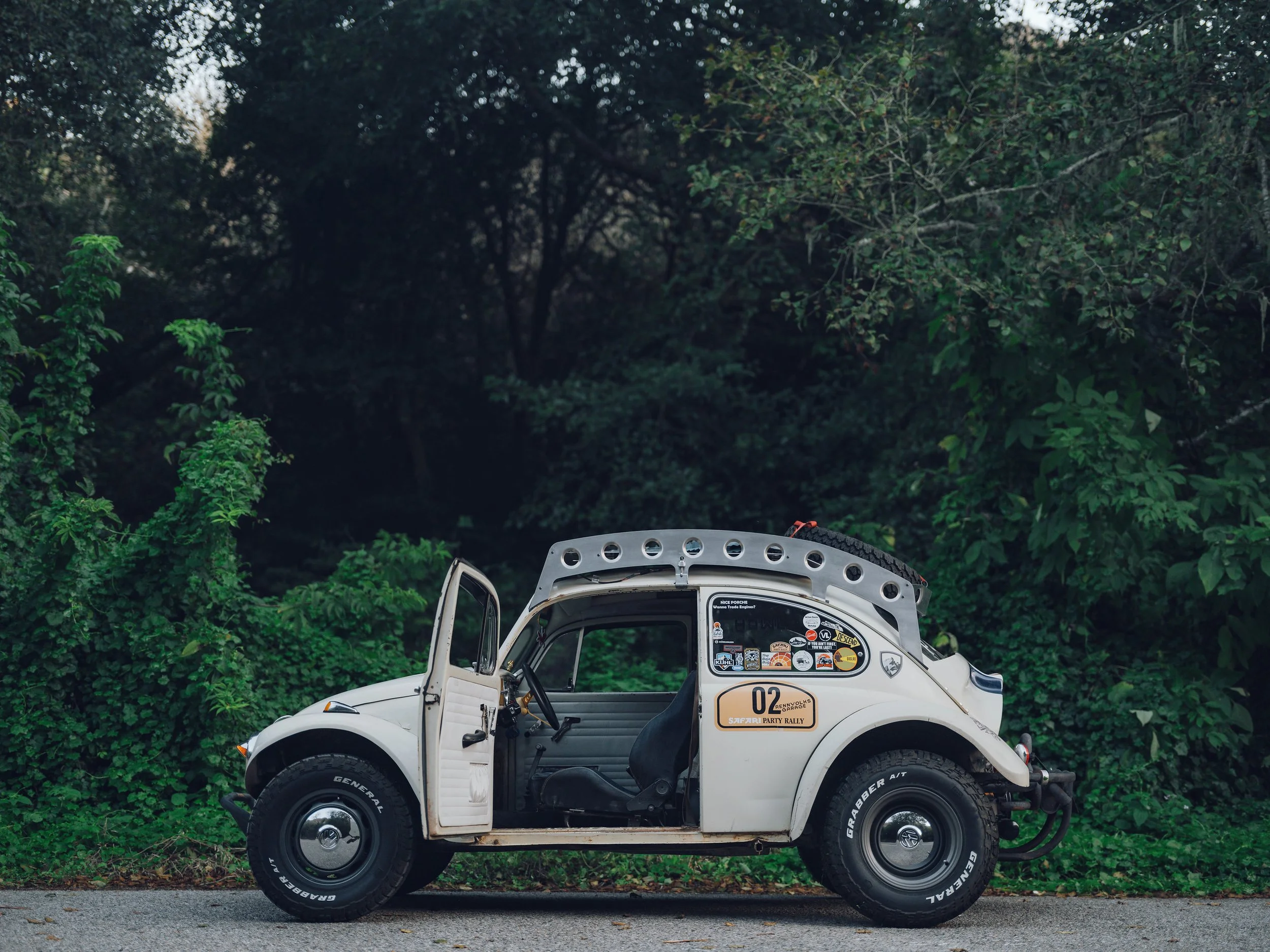 White vintage Volkswagen Beetle with various stickers on the side, parked on the side of a road with green trees and foliage in the background.
